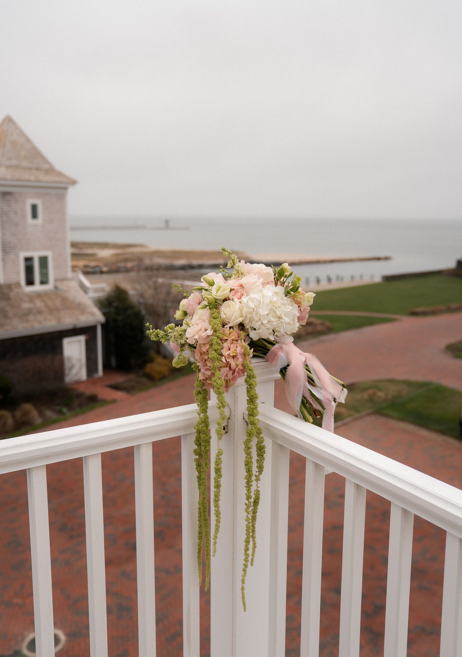 A romantic bridal bouquet of pink and white flowers adorns a white railing overlooking a coastal waterfront venue.