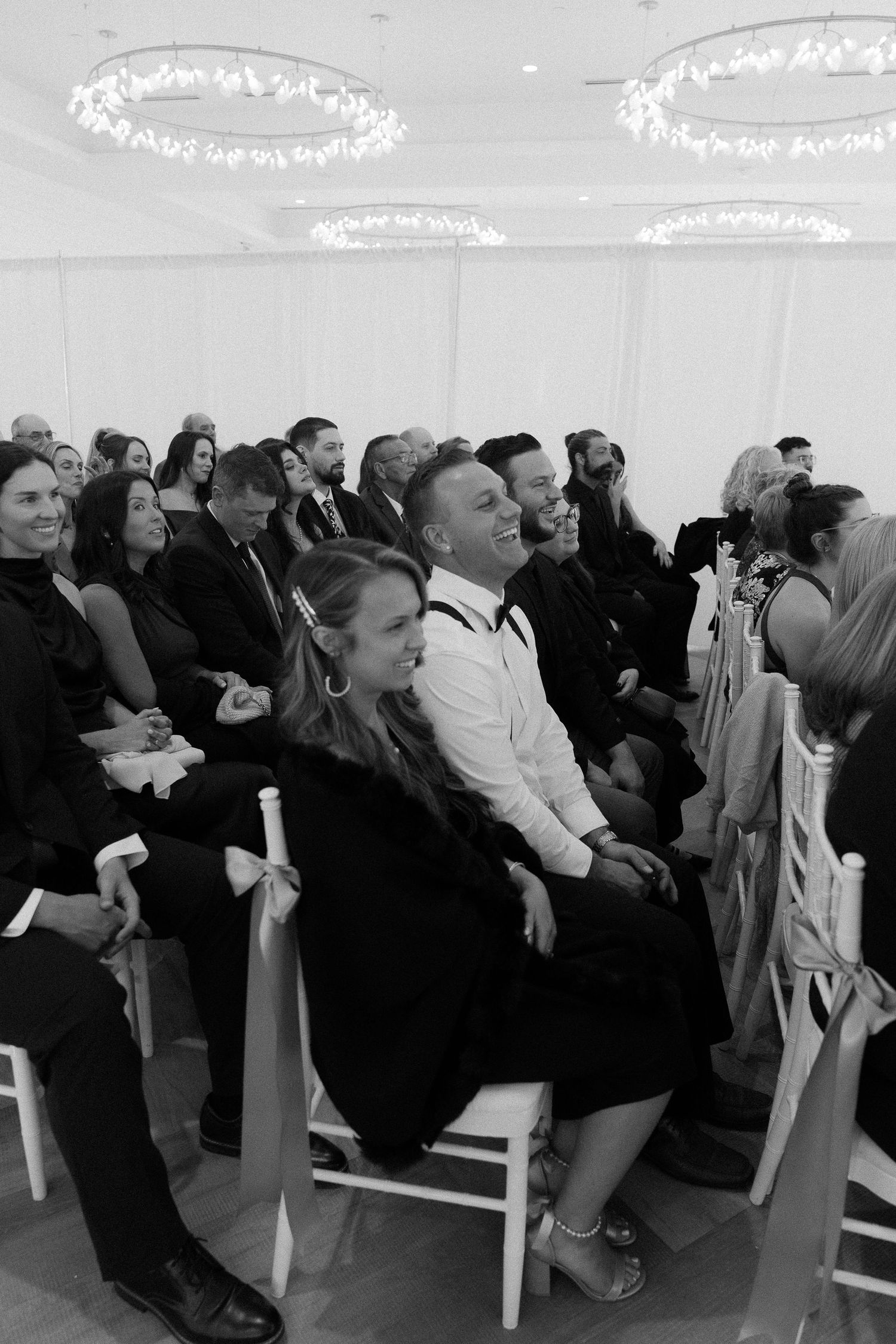 Wedding guests laugh and smile while seated at an indoor ceremony with chandelier lighting in black and white.