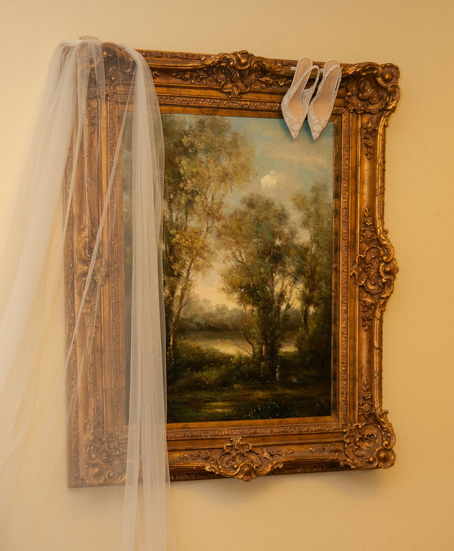 Gold ornate framed landscape painting decorated with a bridal veil and white ribbon bow on a neutral wall.