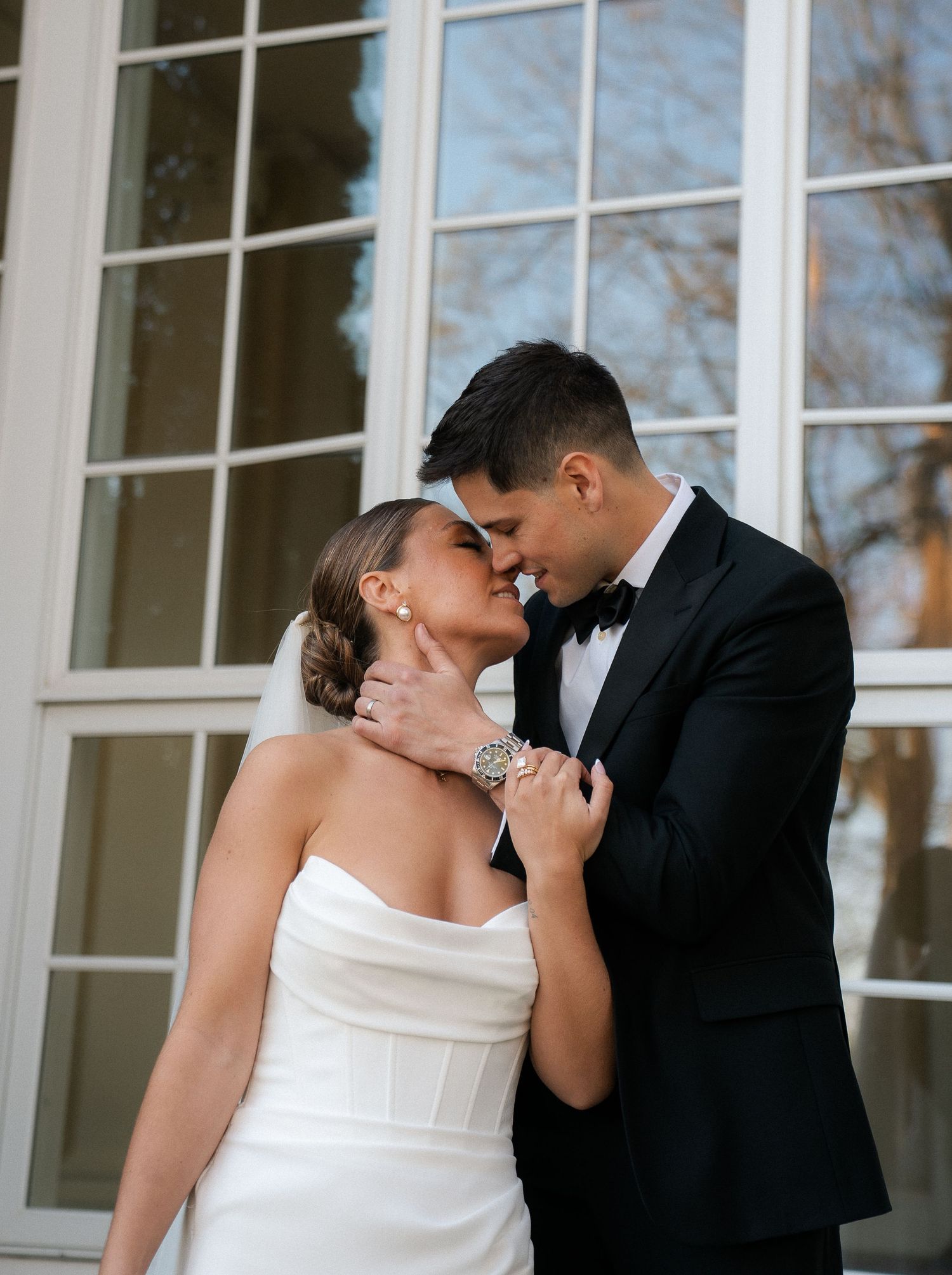 Bride and groom share romantic almost-kiss moment in front of white French windows on wedding day.
