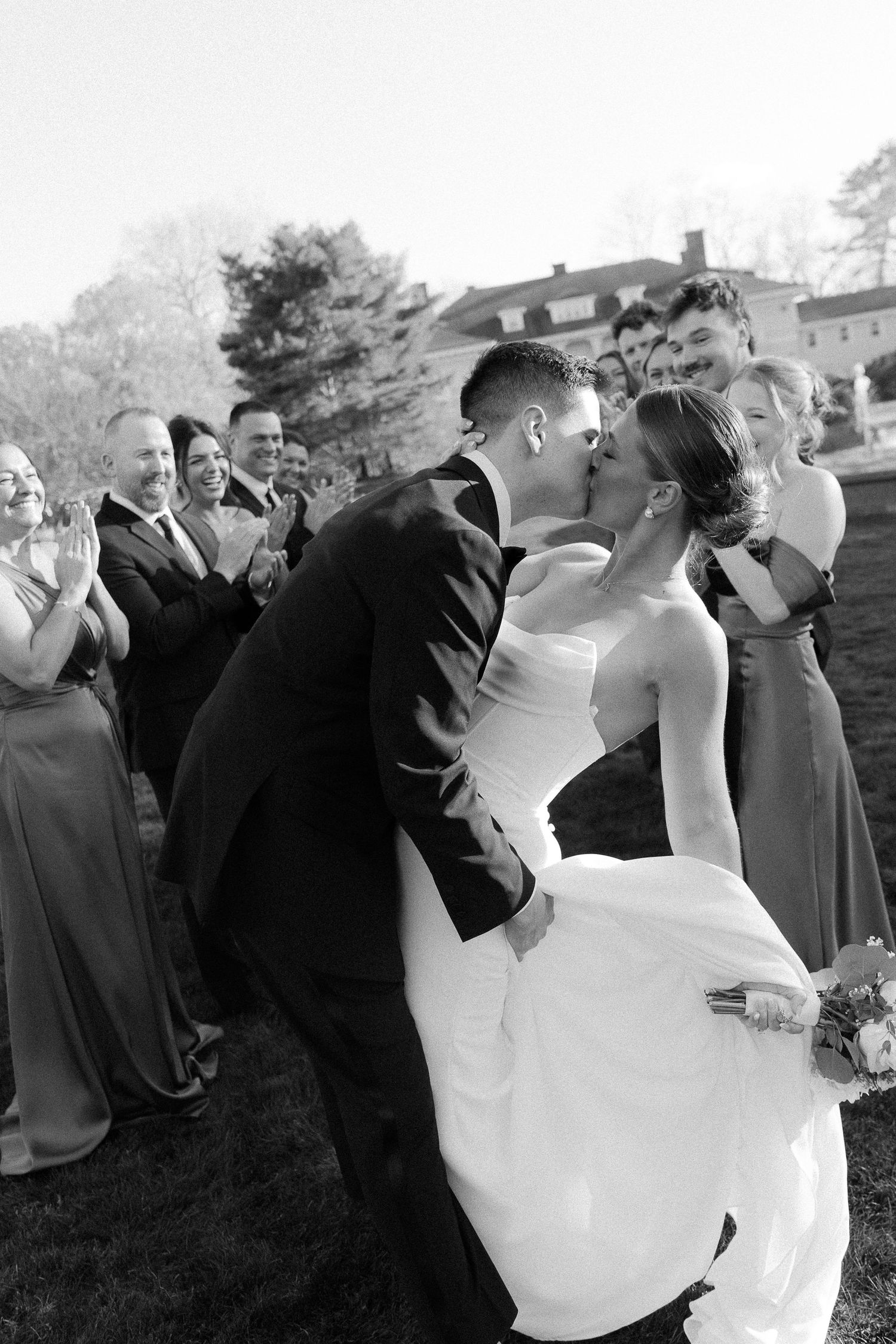 Black and white photo of a bride and groom sharing a kiss surrounded by their wedding party outdoors.