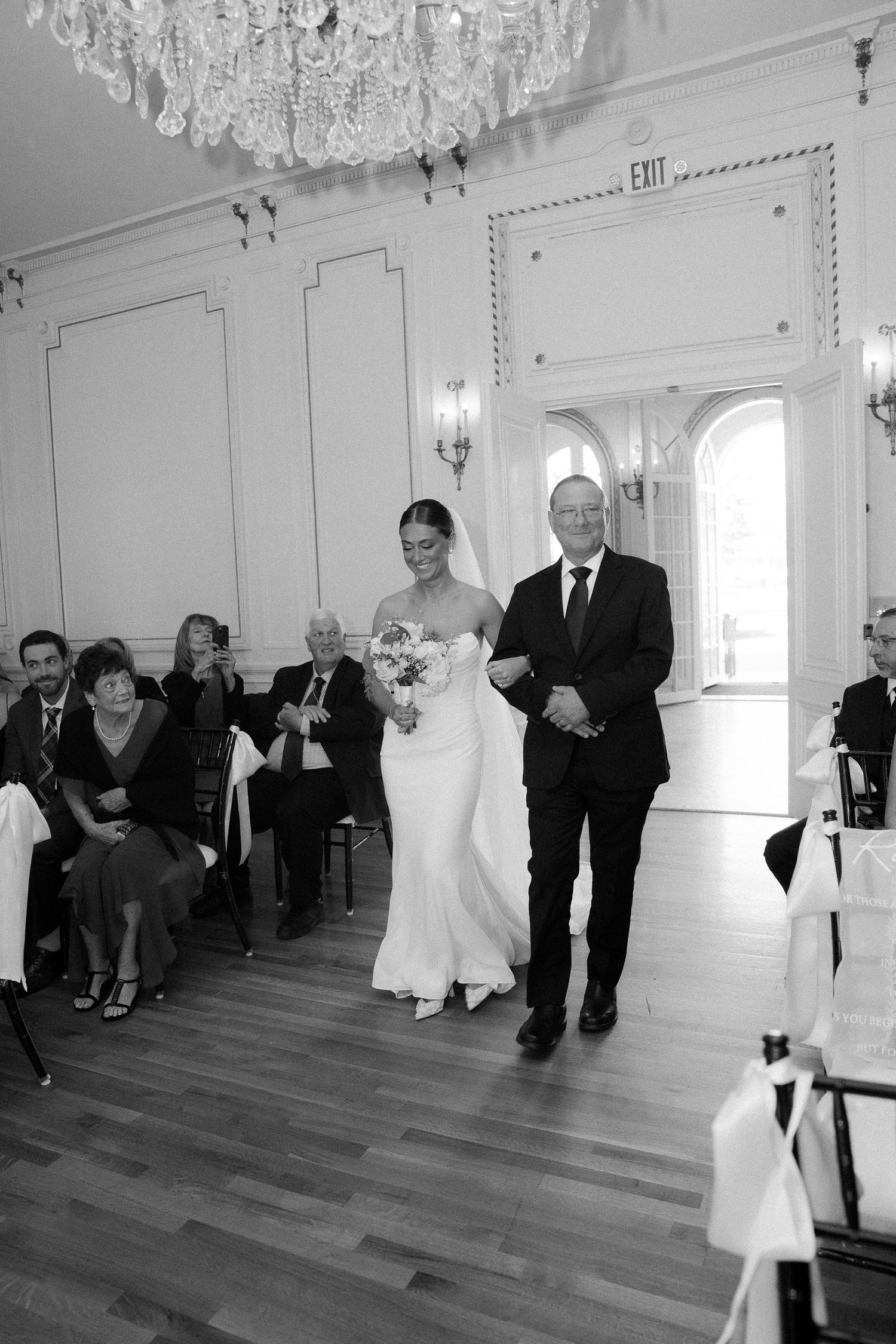 Black and white photo of bride walking down aisle with father in grand ballroom.