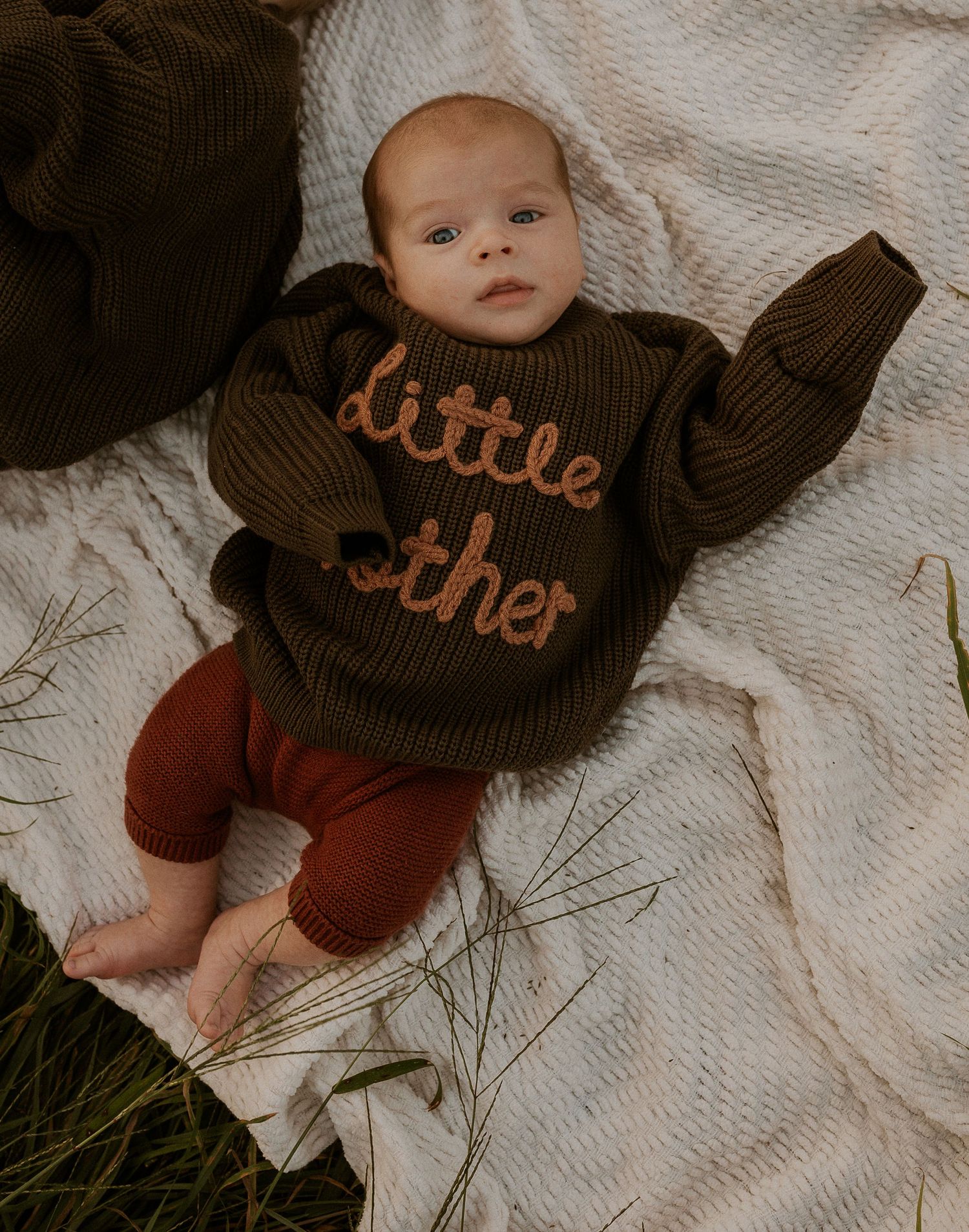 Baby wearing brown 'little brother' sweater lying on white blanket outdoors.