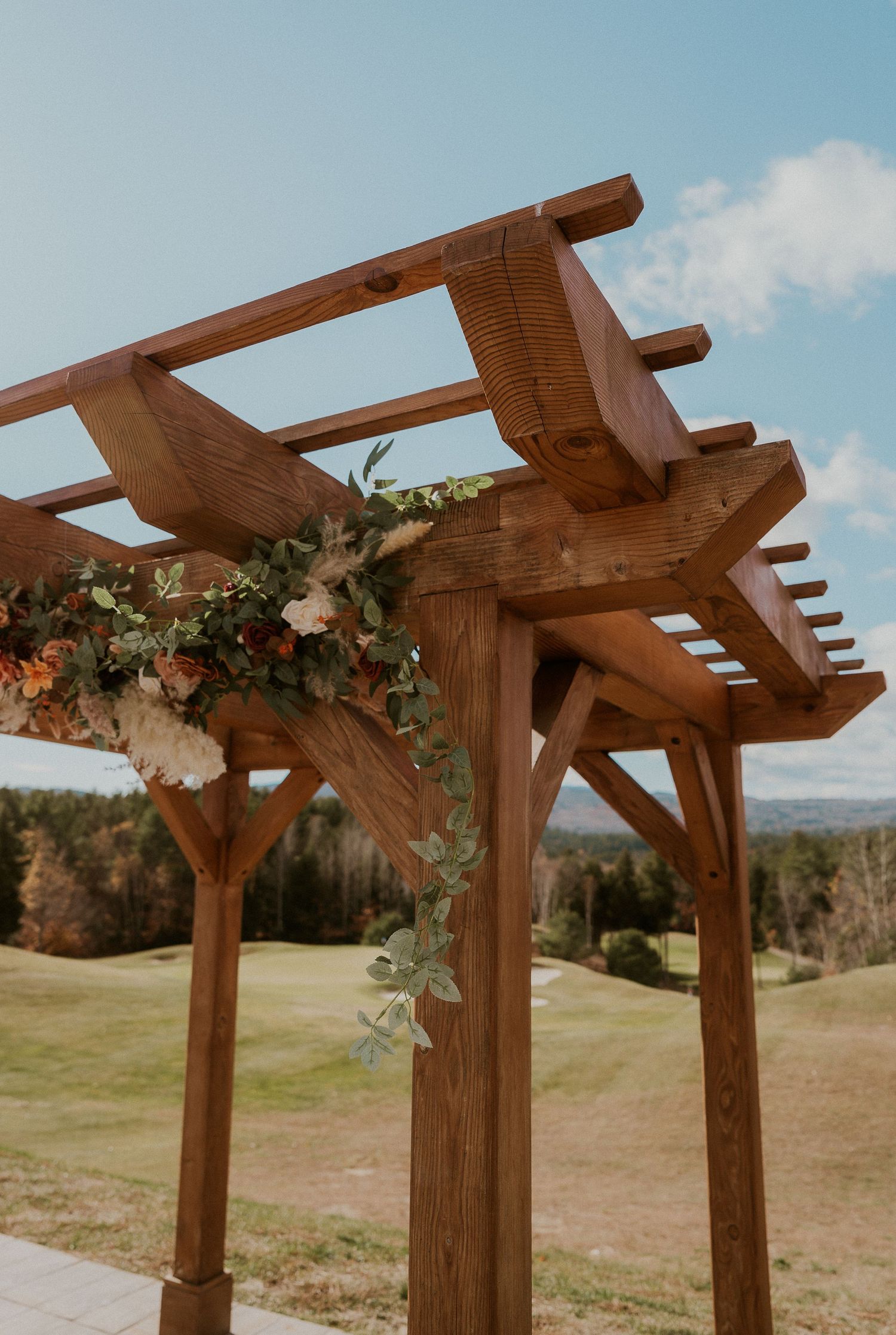 Wooden wedding arbor decorated with peach flowers and greenery overlooks a scenic mountain landscape on a sunny day.