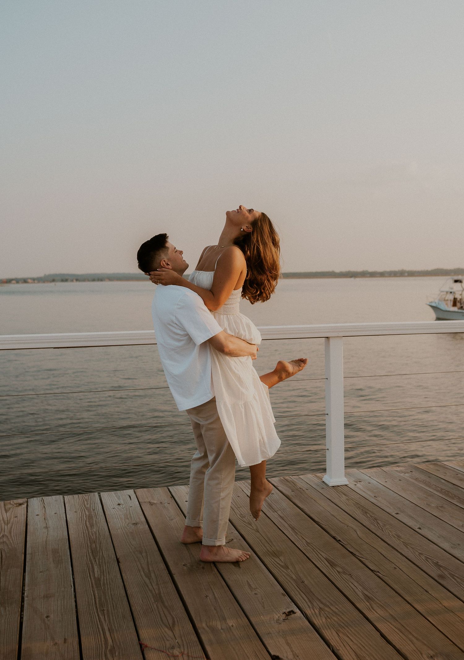 A couple shares a romantic moment on a wooden dock at sunset, with the man lifting the woman in a white dress.