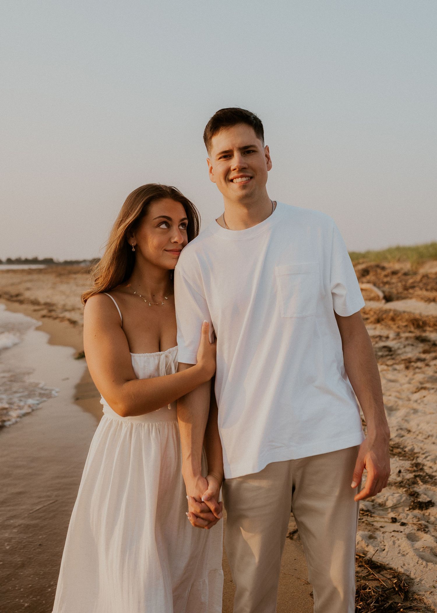 Couple in coordinated white outfits walking hand in hand along a beach shoreline at golden hour.