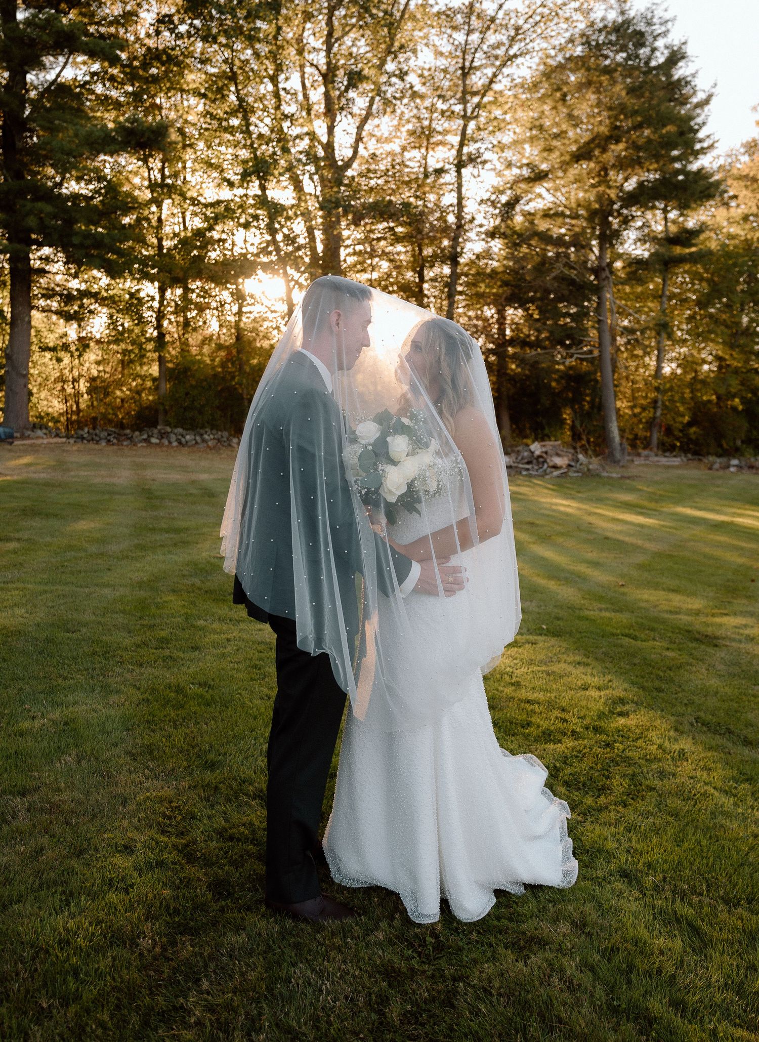 Wedding couple poses in golden hour sunlight with a long flowing veil against a natural outdoor backdrop.