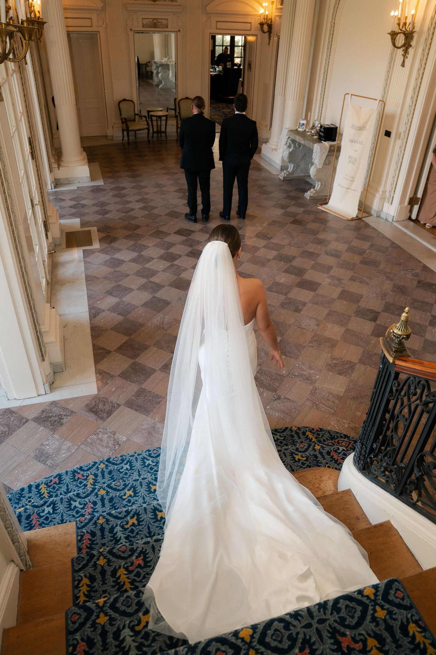 Bride in white gown and veil descends grand staircase toward two groomsmen in elegant wedding venue.