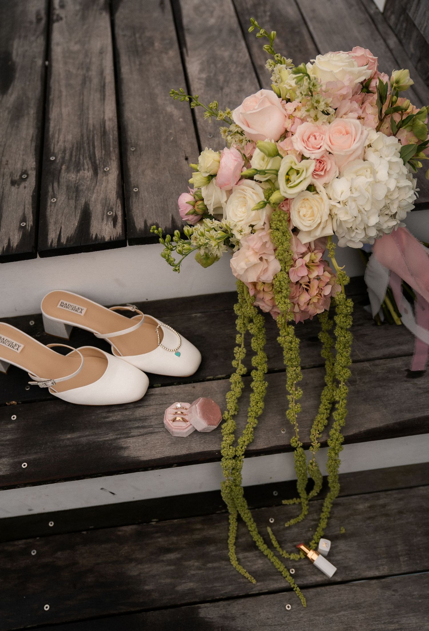 Bridal flat shoes beside a pink velvet ring box and cascading bouquet of blush roses on wooden steps.