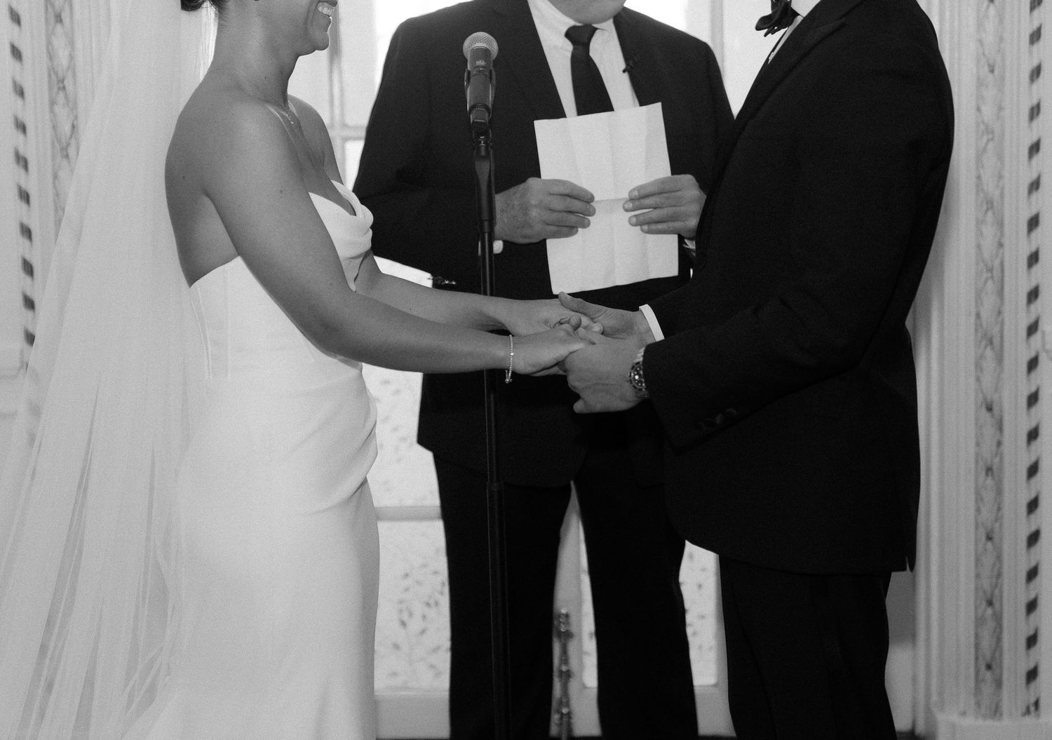 Black and white wedding ceremony photo of bride and groom exchanging vows with officiant at elegant indoor venue.