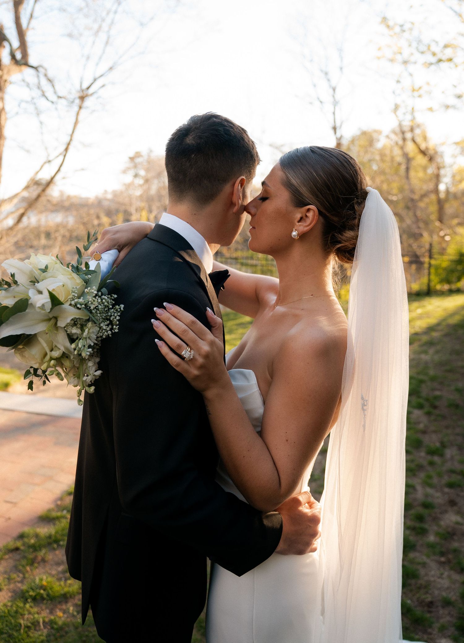 Bride and groom sharing a kiss outdoors, bride in sleek gown with veil holding white floral bouquet.