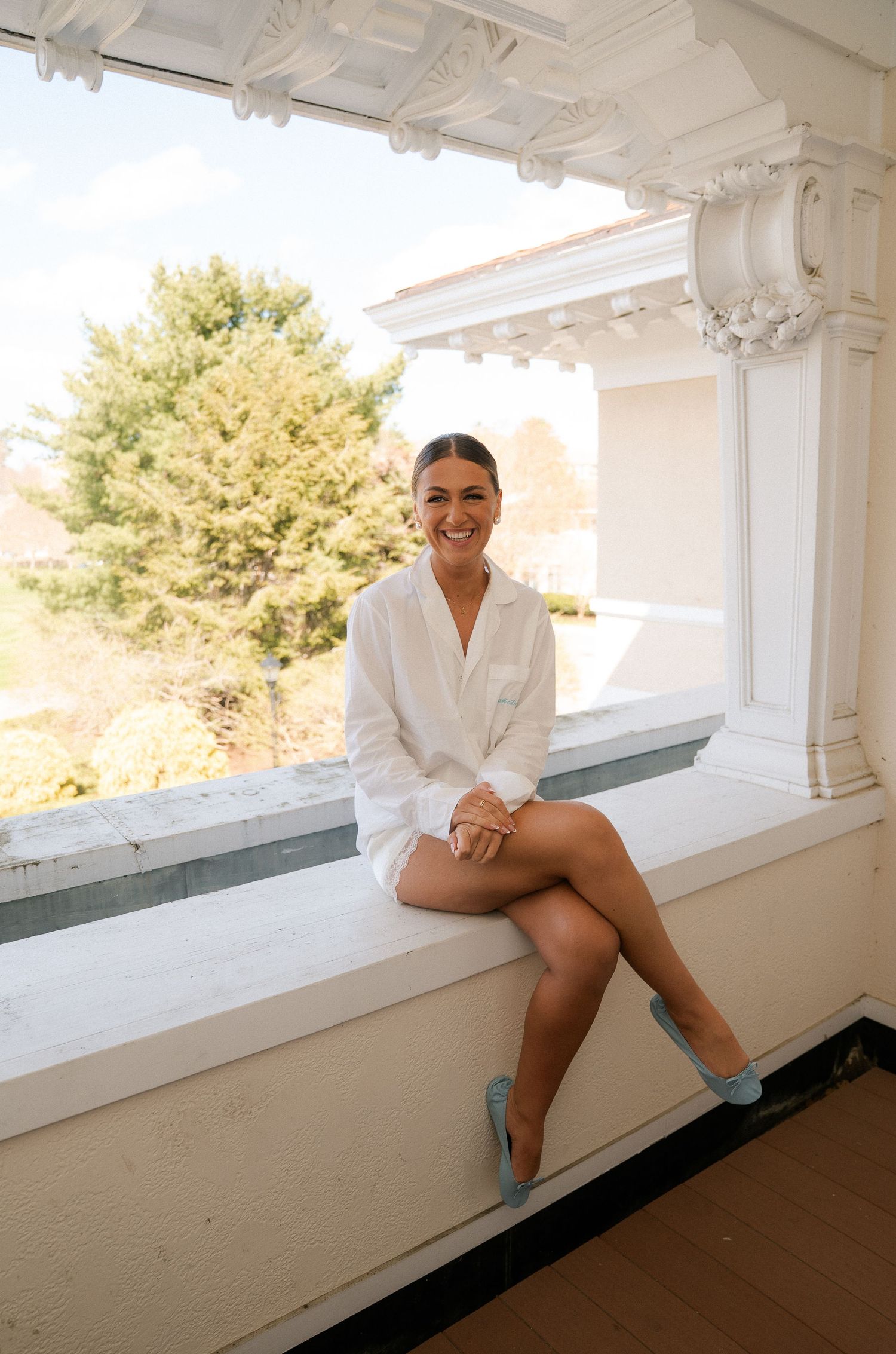 Smiling woman in white dress and blue heels sitting on a white window ledge with trees in the background.