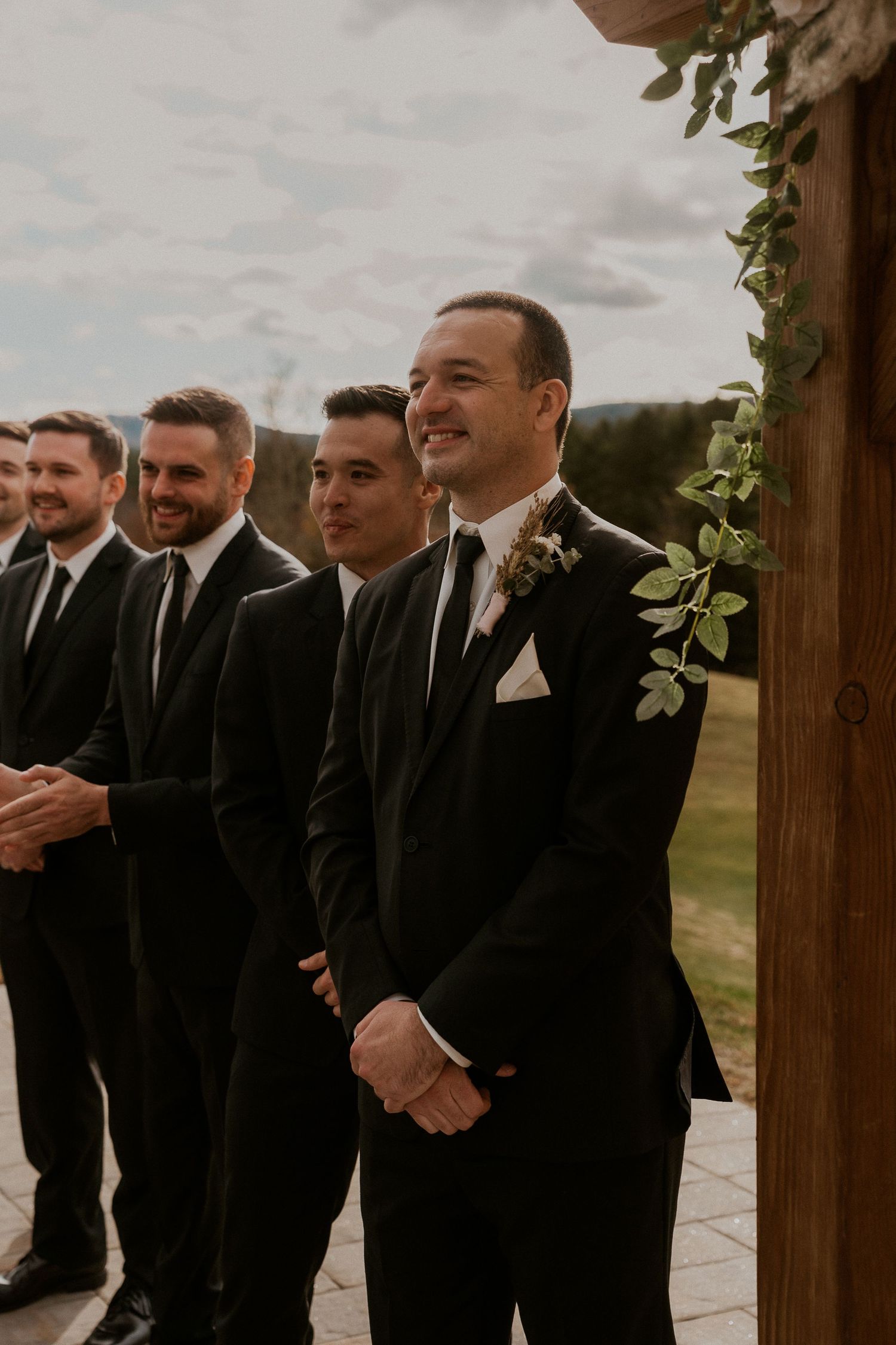 Groomsmen in black suits standing in a line outdoors, sharing smiles and laughter during a wedding ceremony.