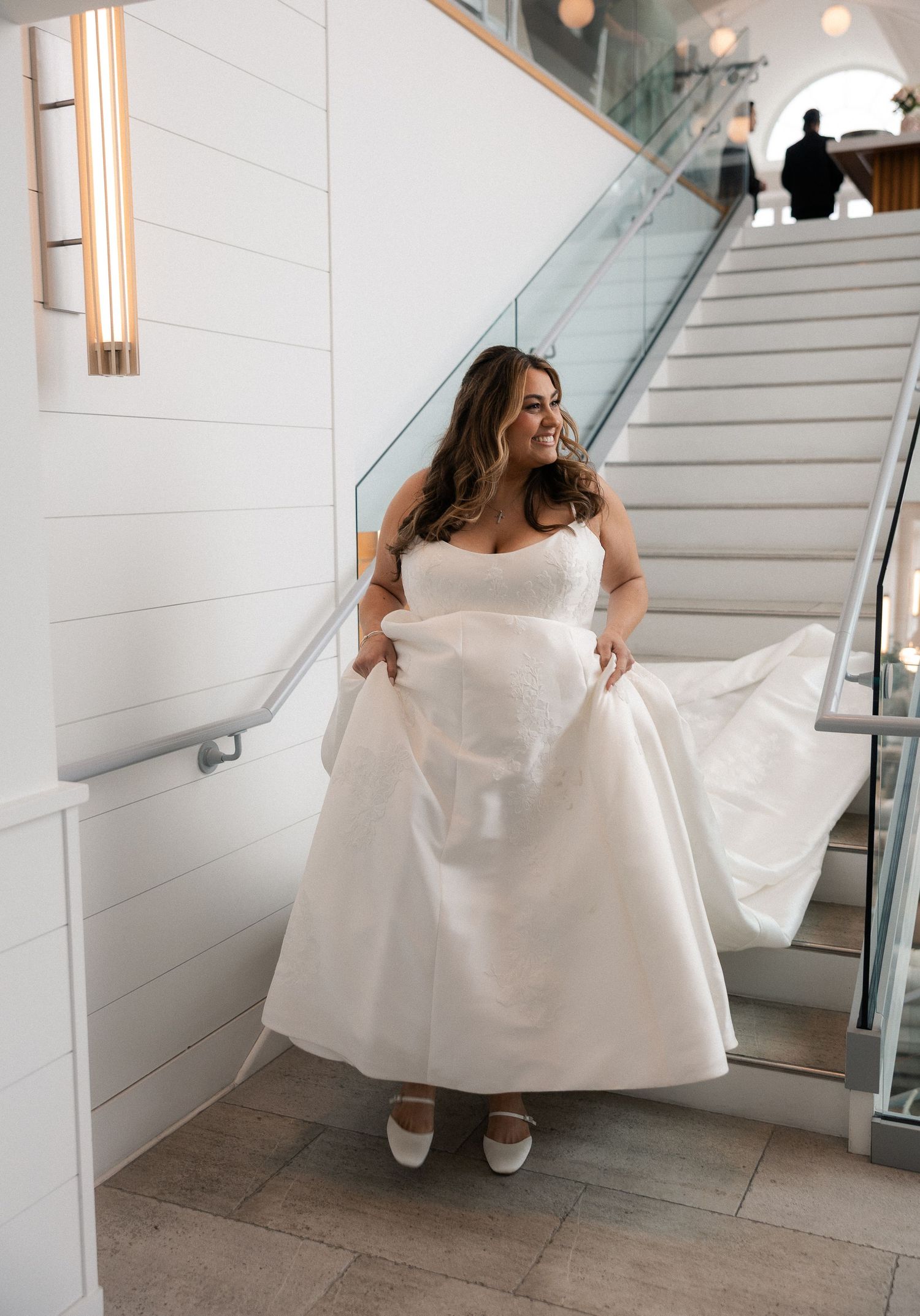 Smiling bride in a strapless white ball gown lifting her skirt while standing near a modern staircase.