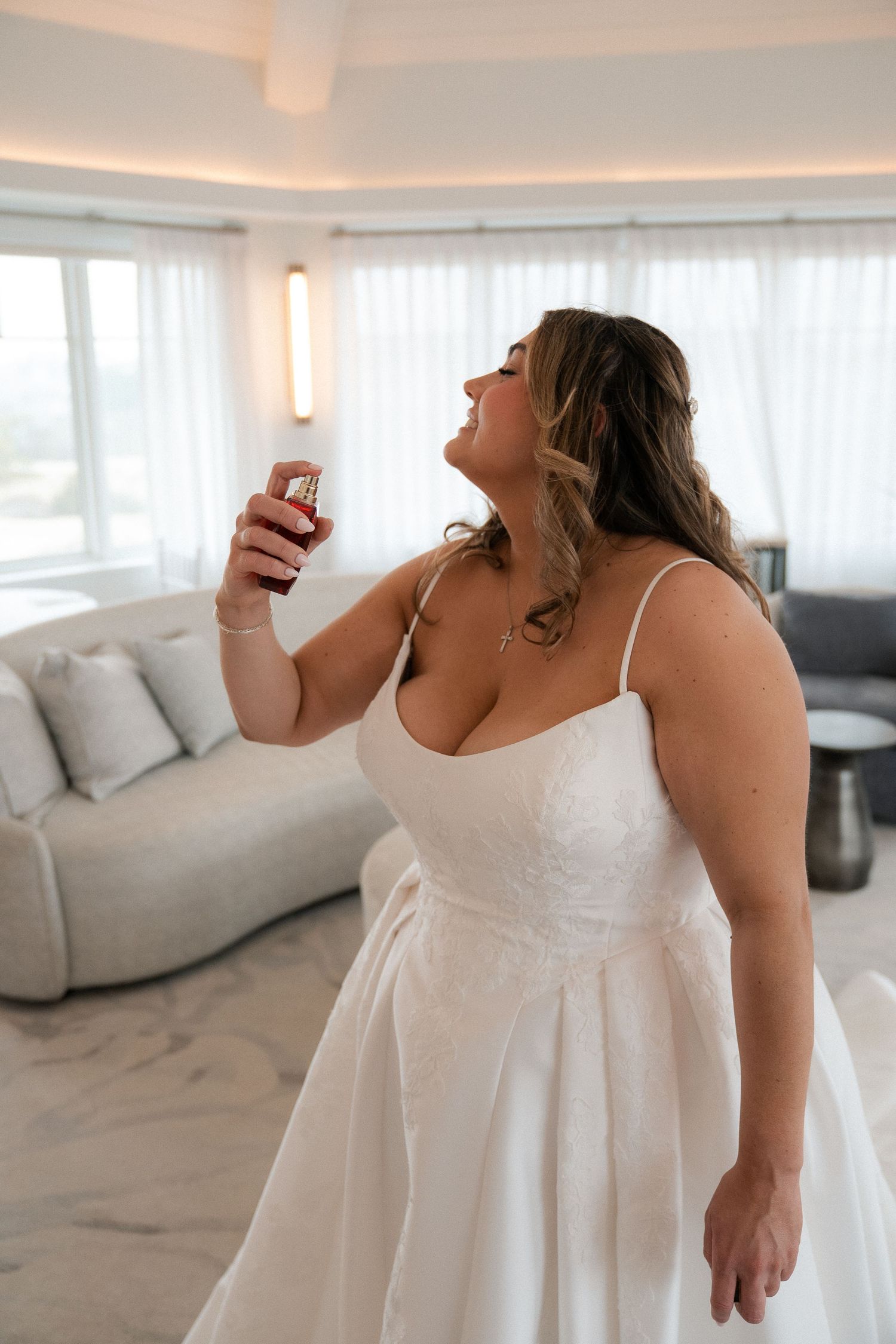 Bride in a white lace wedding gown laughs while holding a perfume bottle in a bright hotel suite.