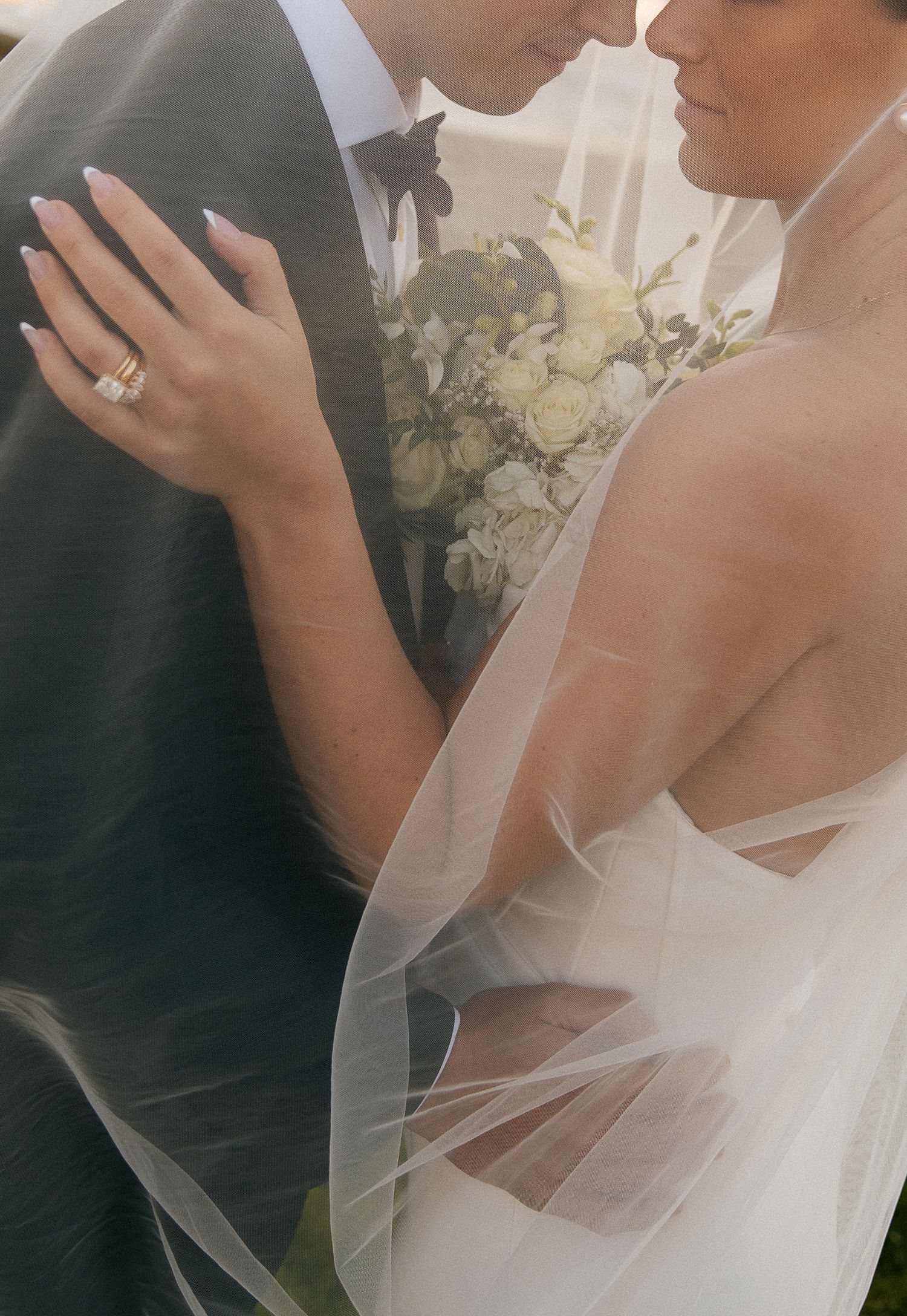 Close-up of bride holding white floral bouquet embracing groom under sheer wedding veil.