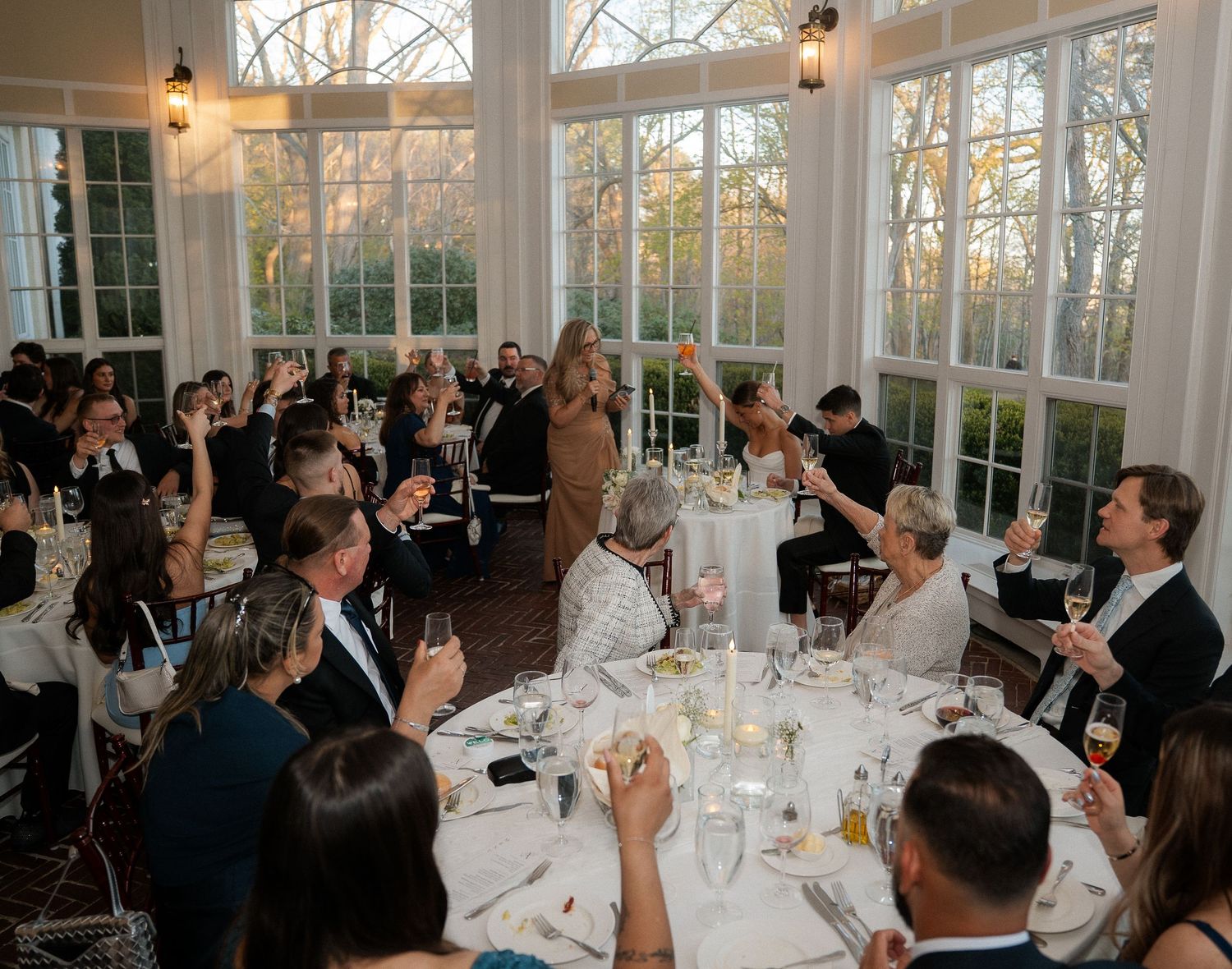 Elegant wedding reception toast with guests raising glasses in a sunlit conservatory dining room.