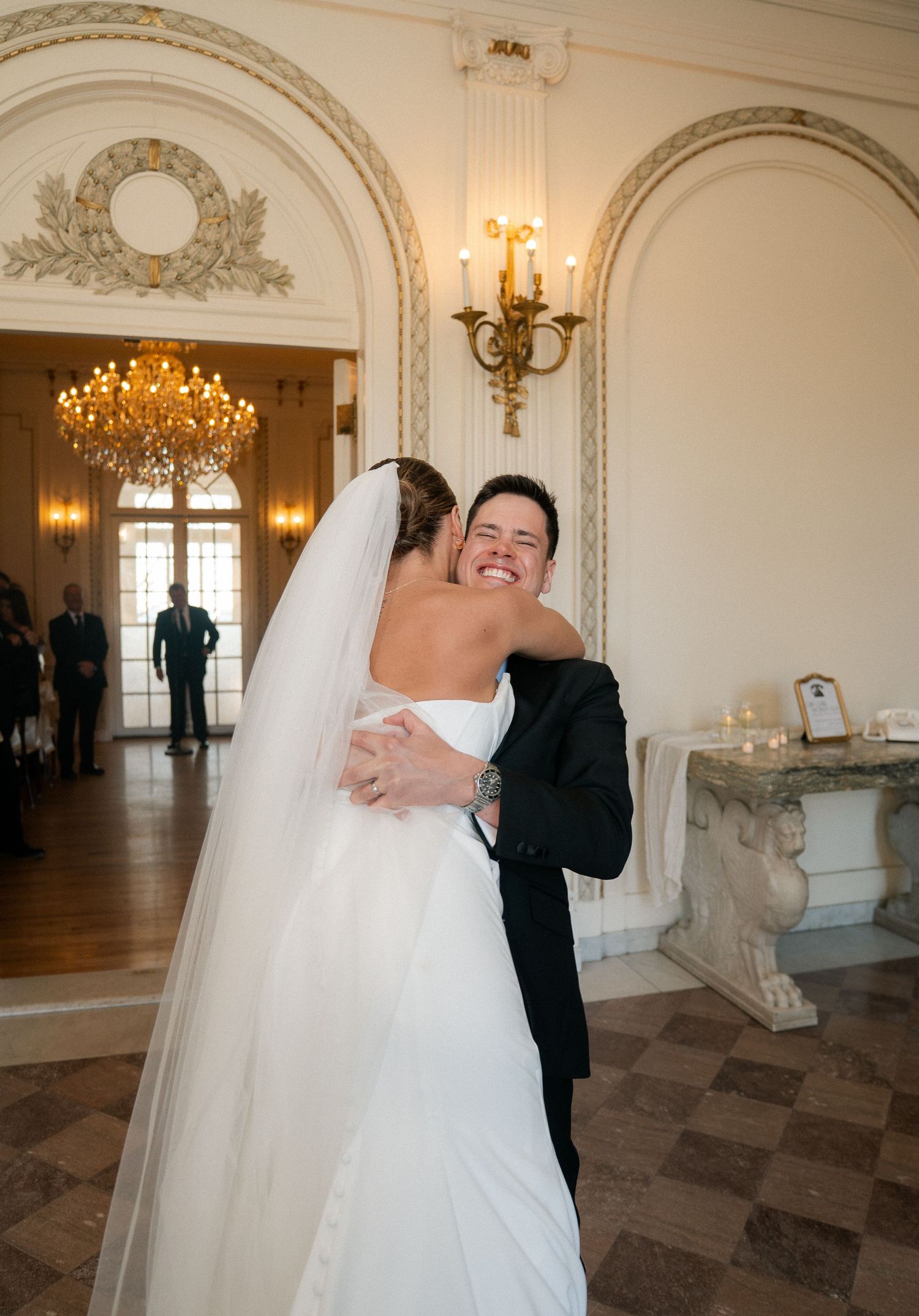 Bride and groom share a joyful embrace in an elegant ballroom with chandelier and arched windows.
