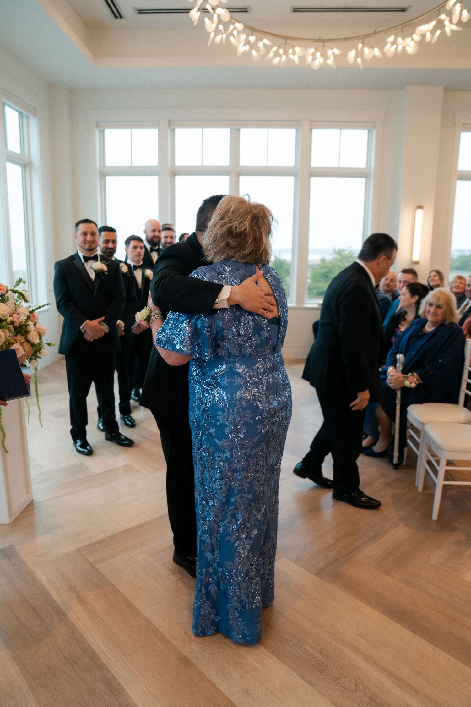 A woman in a blue lace gown embraces a groom during a wedding ceremony in an elegant venue.
