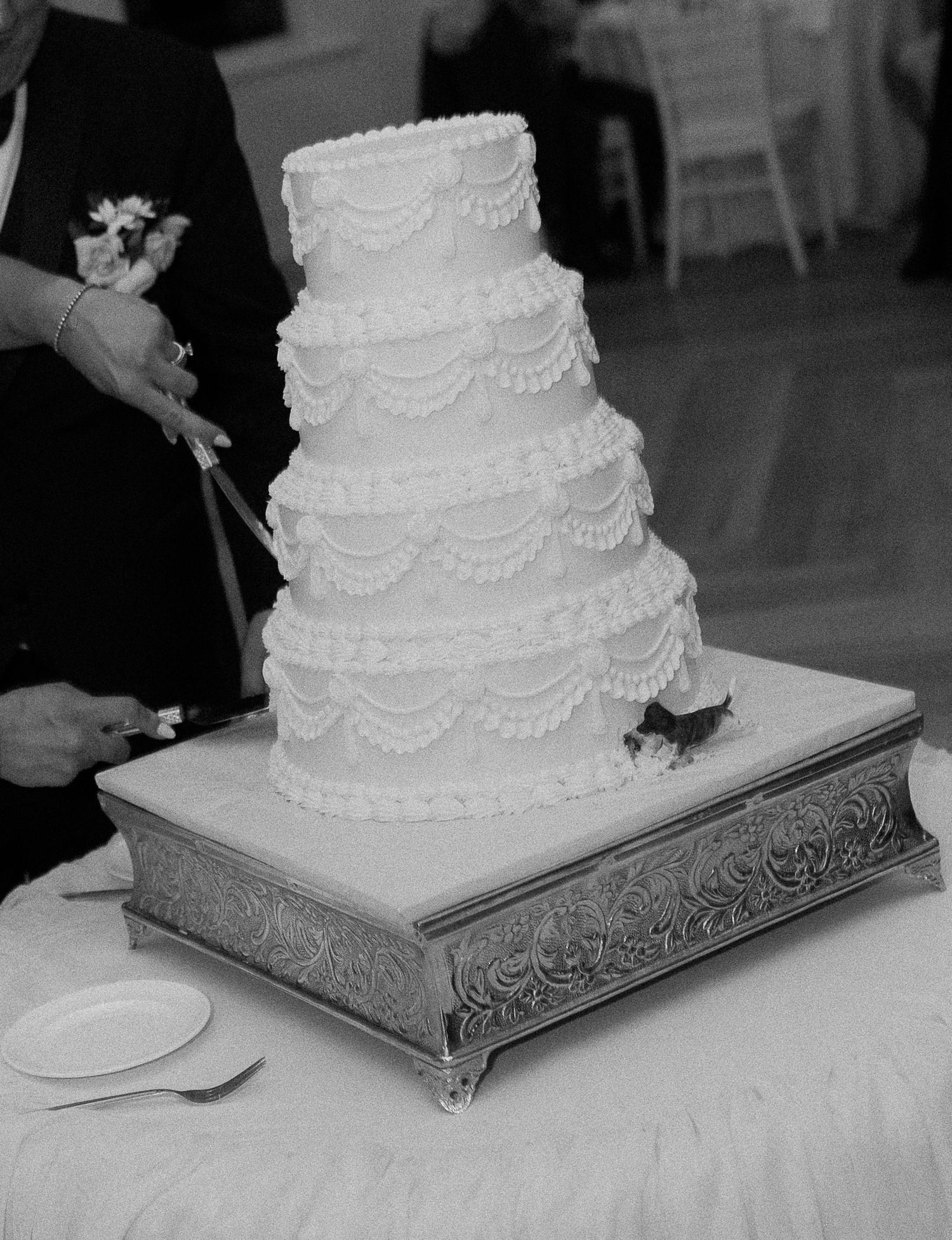 Elegant four-tier white wedding cake with ruffled frosting on ornate silver stand being cut by couple.