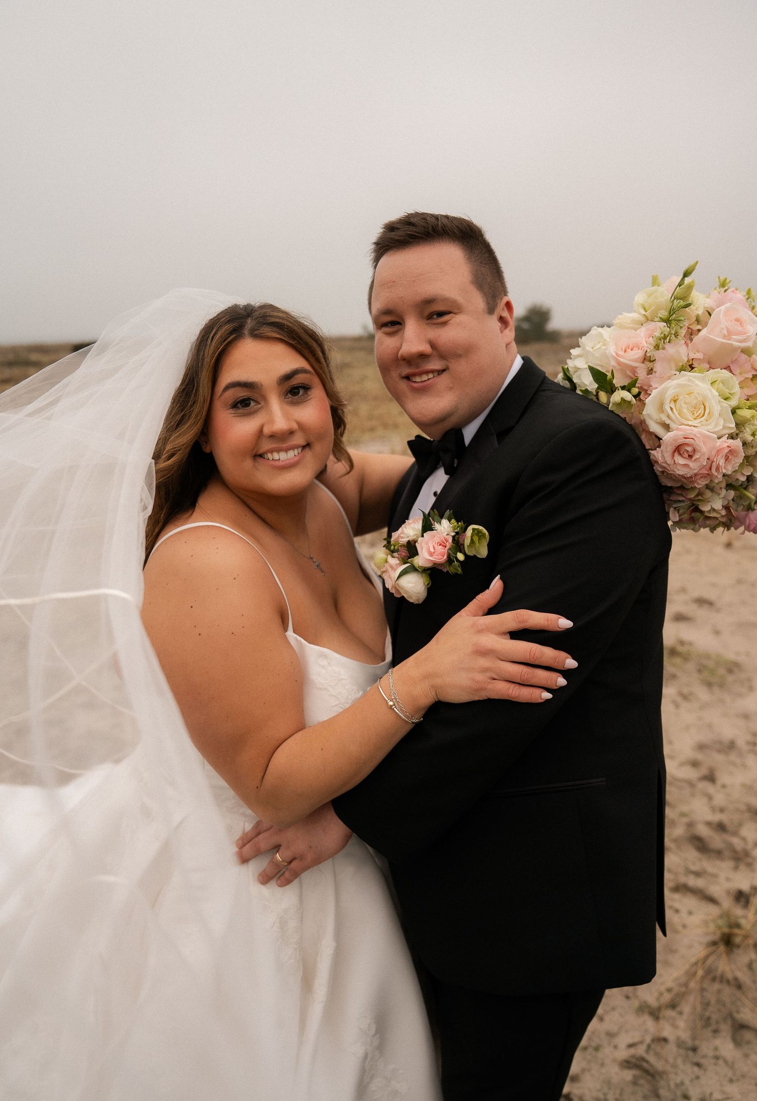 Bride and groom smiling together on sandy beach, bride in white gown with flowing veil.