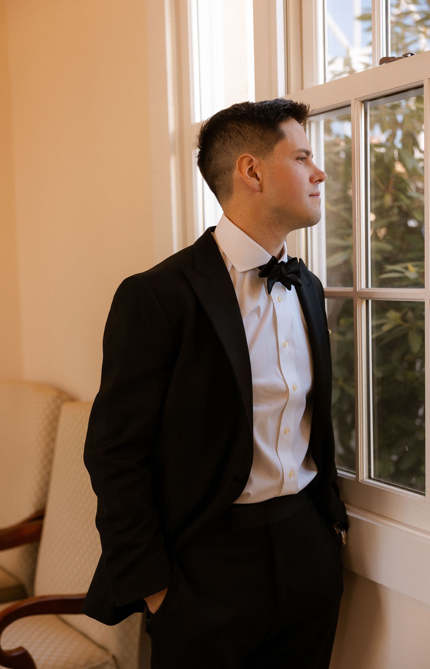 Elegant groom in black tuxedo and bow tie gazing out a window, perfect for wedding inspiration.