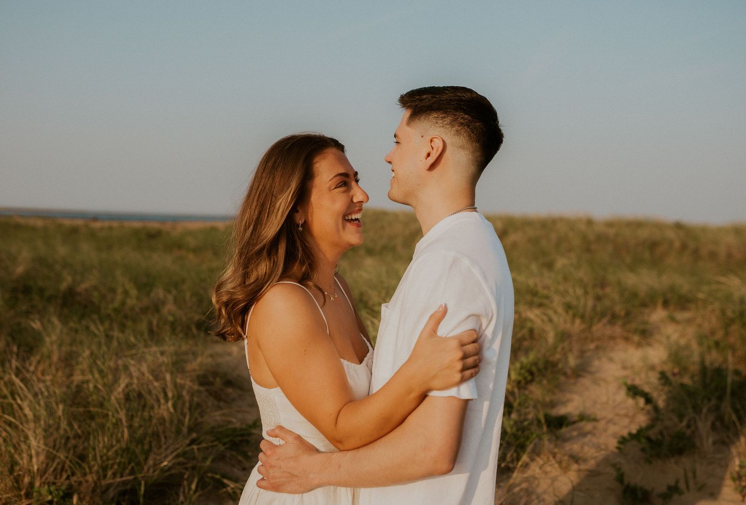 Golden hour photos of a couple embracing on a sandy path with prairie grass in the background.