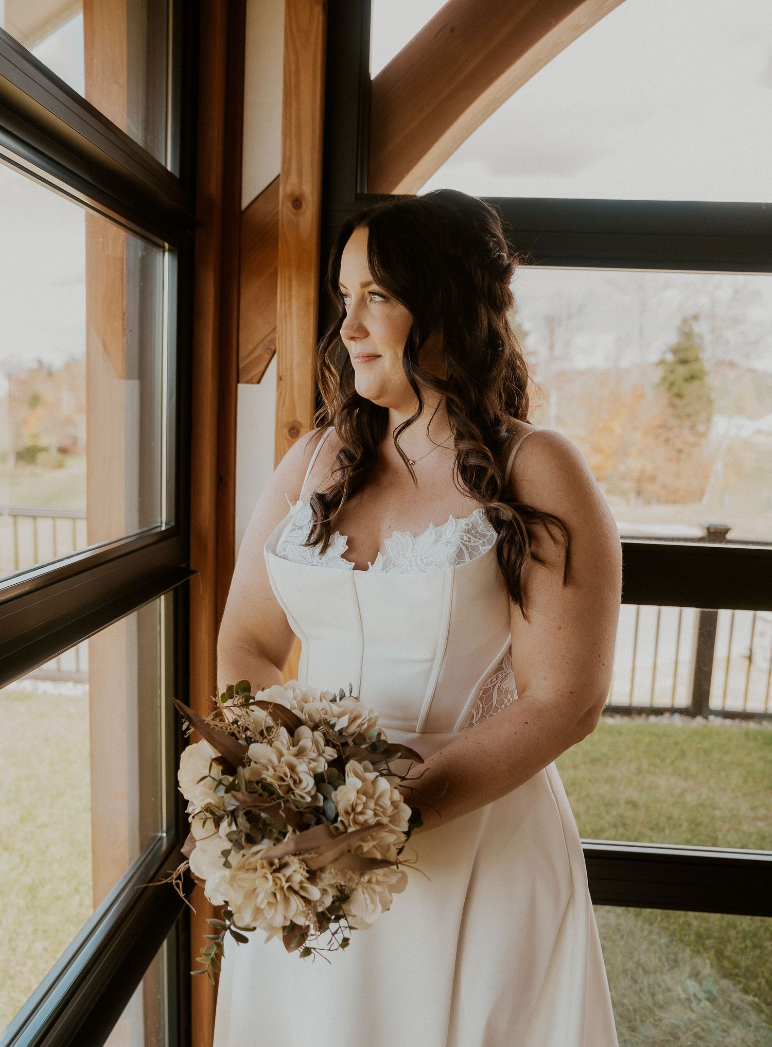 A bride in a white lace dress gazes out a window while holding a neutral-toned bouquet in warm sunlight.
