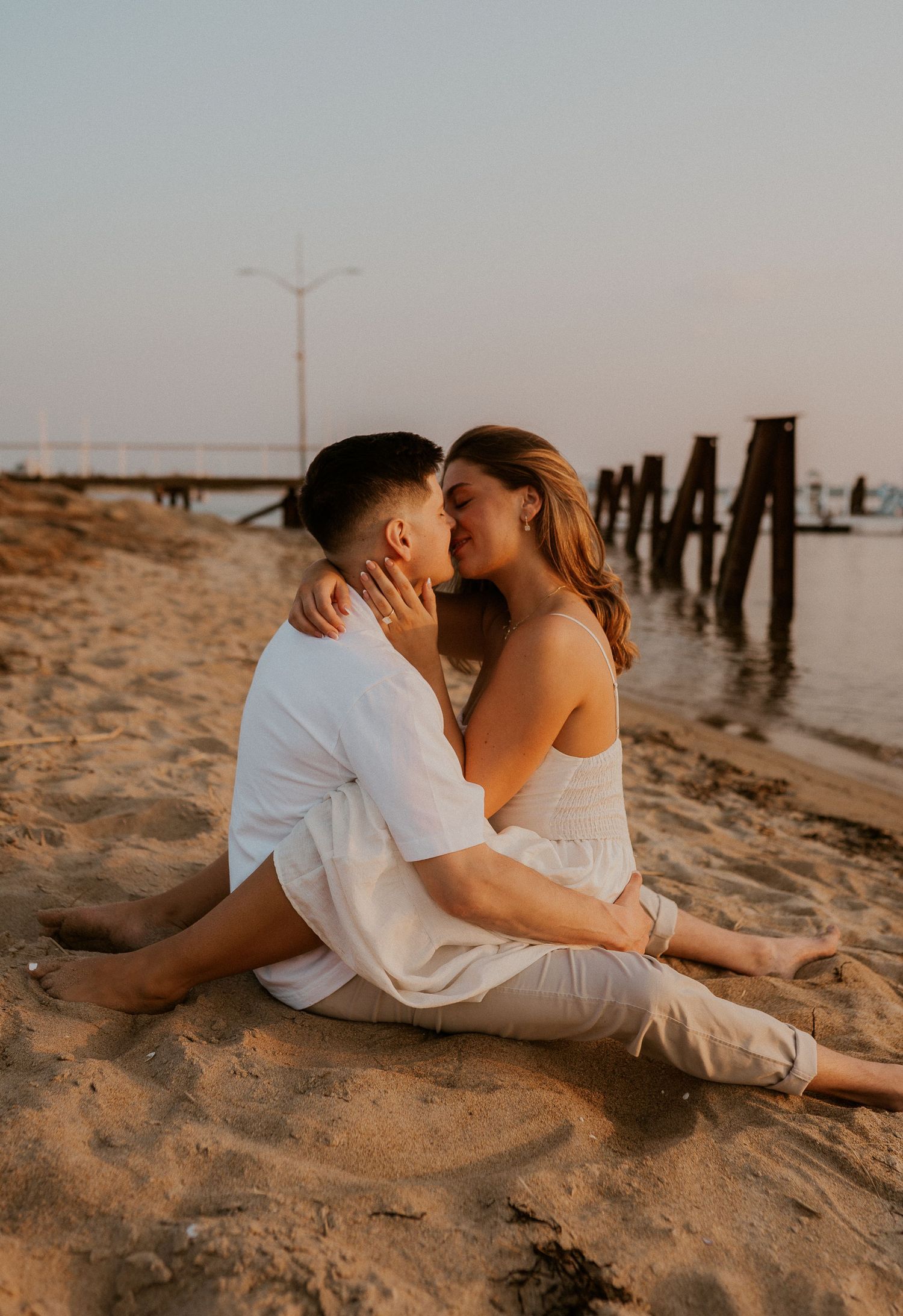 Intimate moment between couple sitting on beach with pier posts silhouetted against sunset sky.