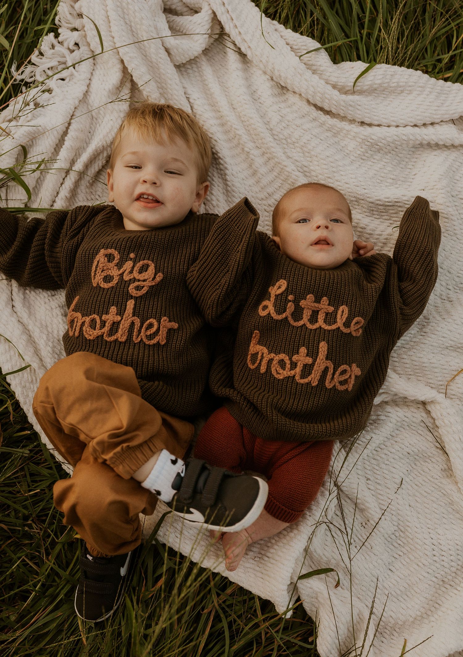 Two siblings wearing matching brown sweaters lying on white blanket outdoors.