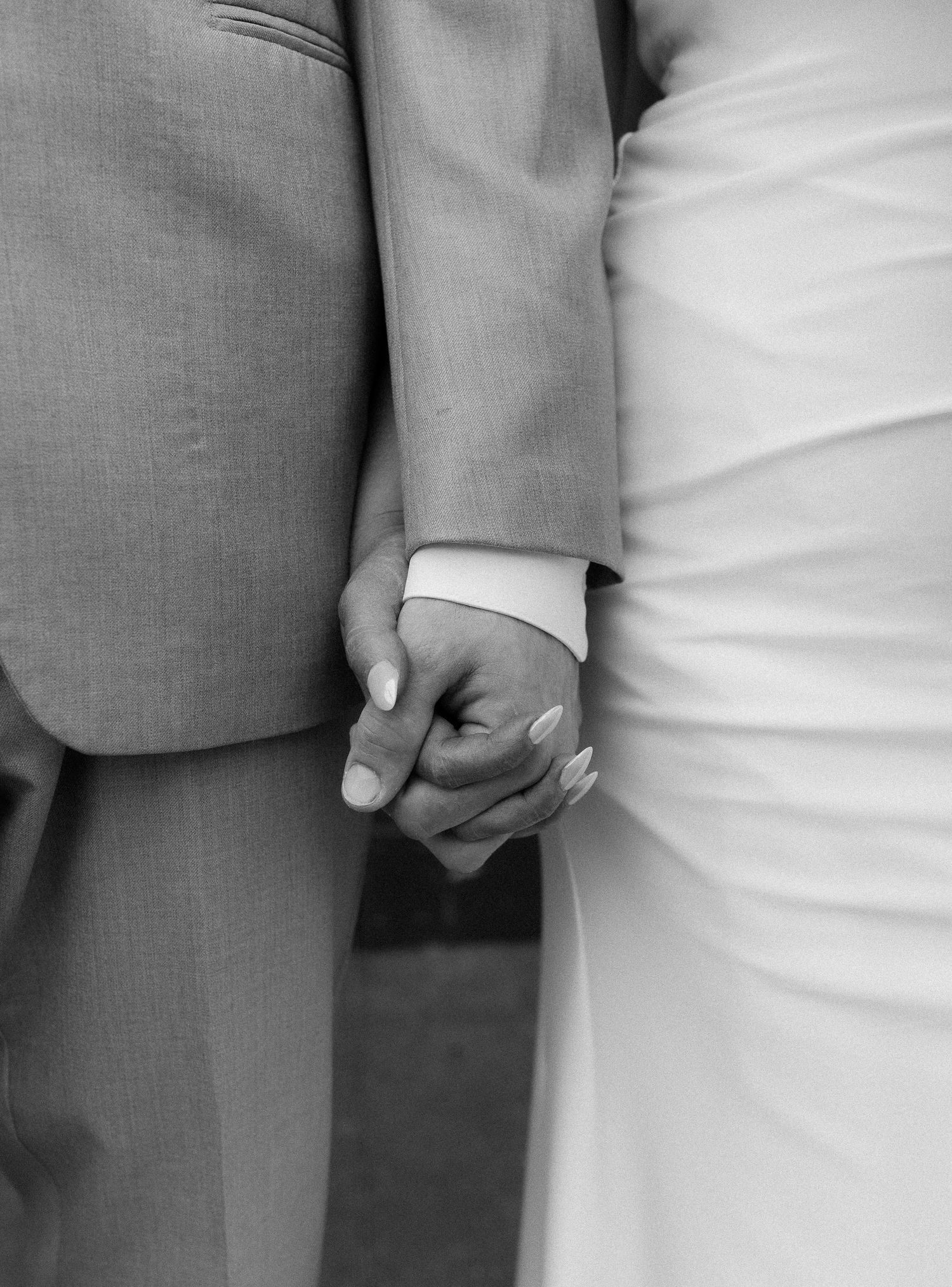 Close-up of couple holding hands showing wedding attire details in black and white.