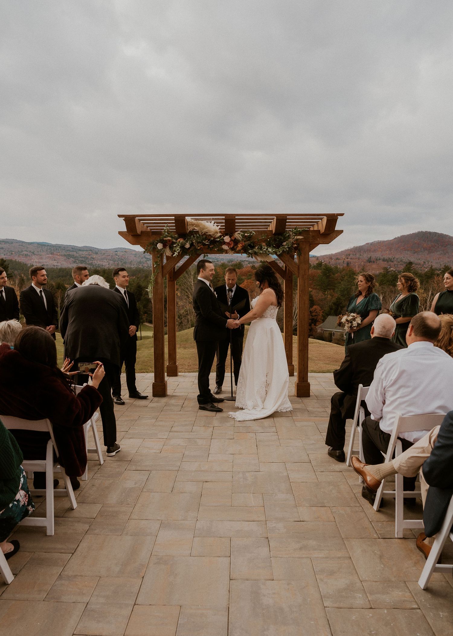 Wedding ceremony taking place on an outdoor stone patio with wooden pergola and mountain views in the background.