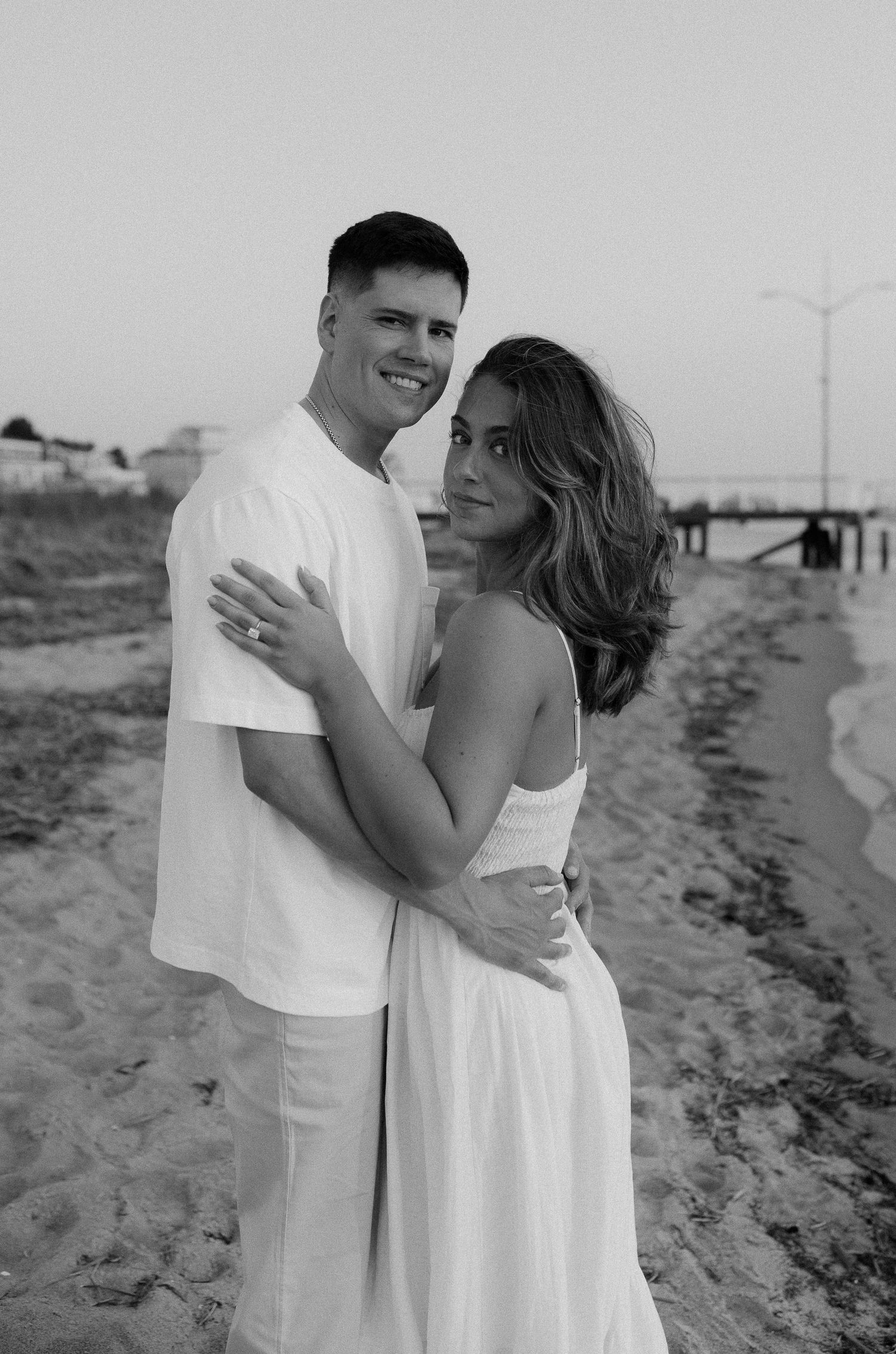 A romantic couple in white outfits embraces on a sandy beach in black and white photography.
