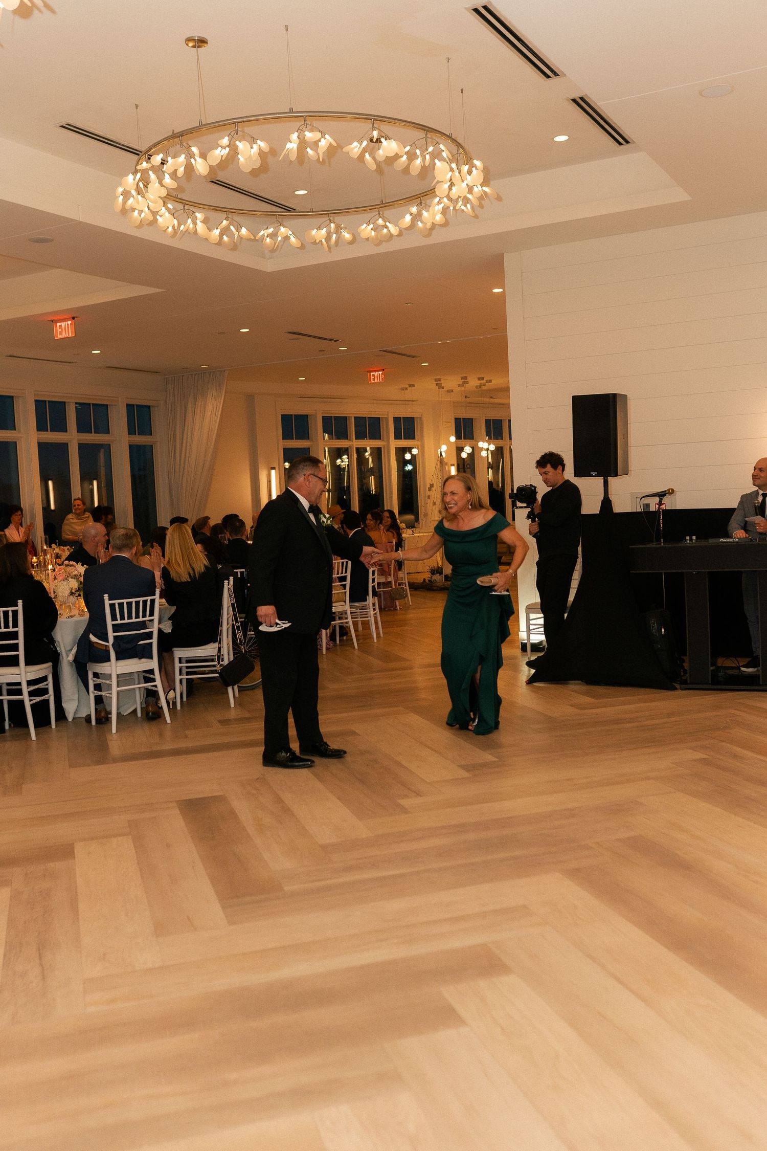 Couple shares a dance on elegant herringbone wood floor beneath floral chandelier at upscale wedding reception.