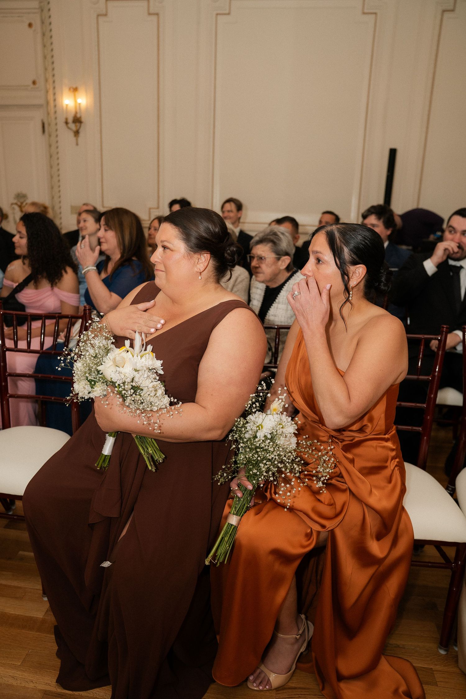 Two emotional bridesmaids in brown and copper satin gowns holding white bouquets during a wedding ceremony.