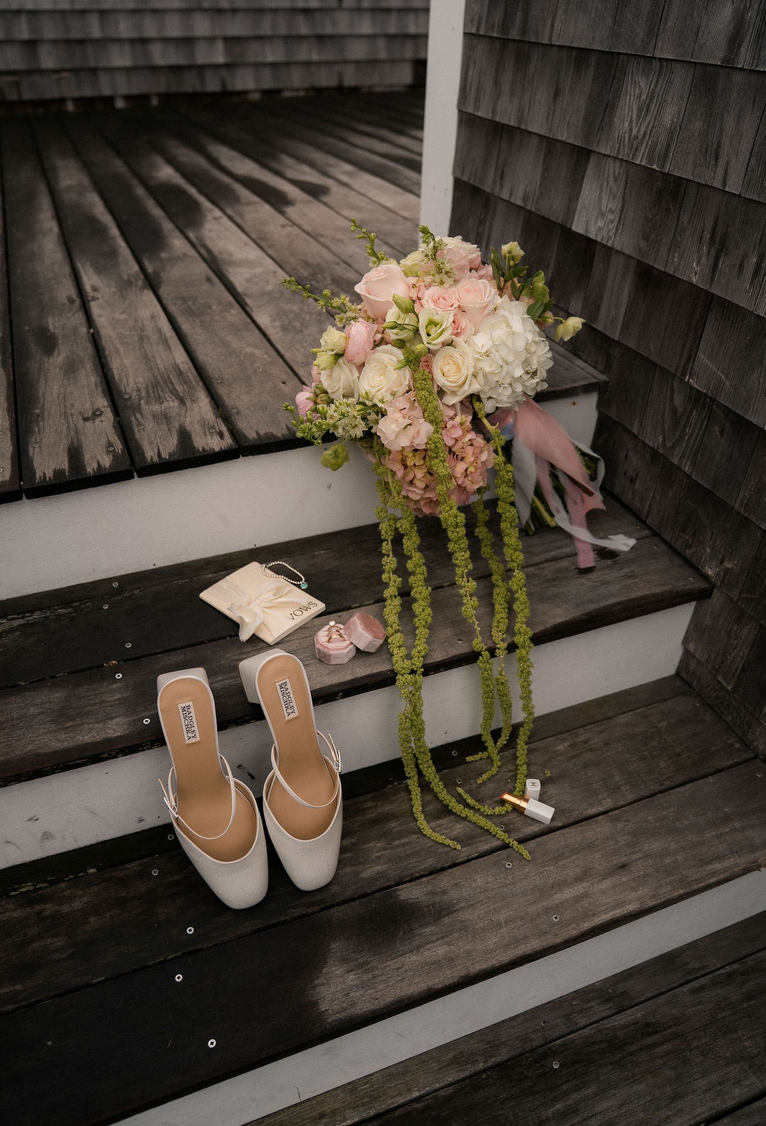 Bridal bouquet of pink roses and hydrangeas with trailing greenery on wooden steps beside white mule heels.
