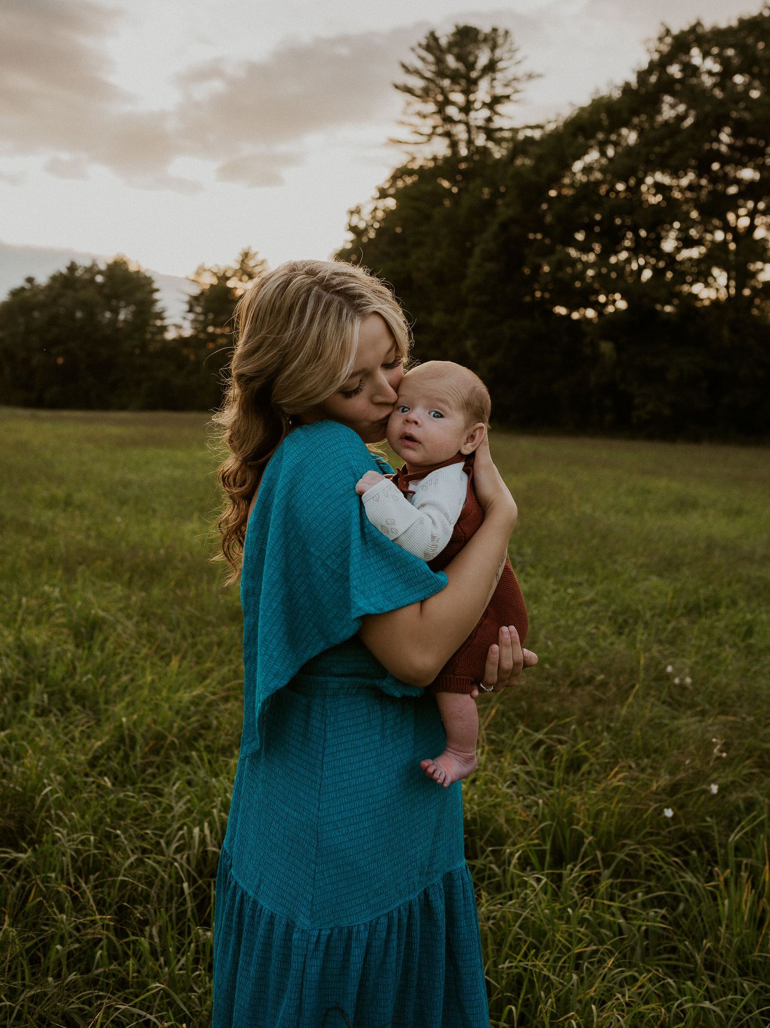 Woman in teal dress tenderly holds infant in sunset-lit meadow.