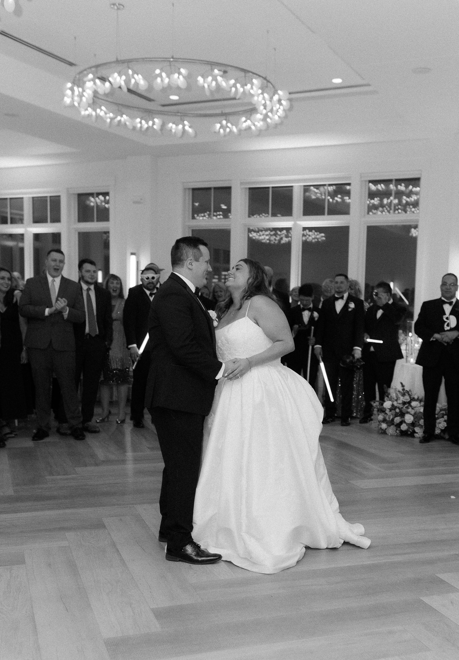 Bride and groom share first dance under chandelier while guests applaud in elegant ballroom.