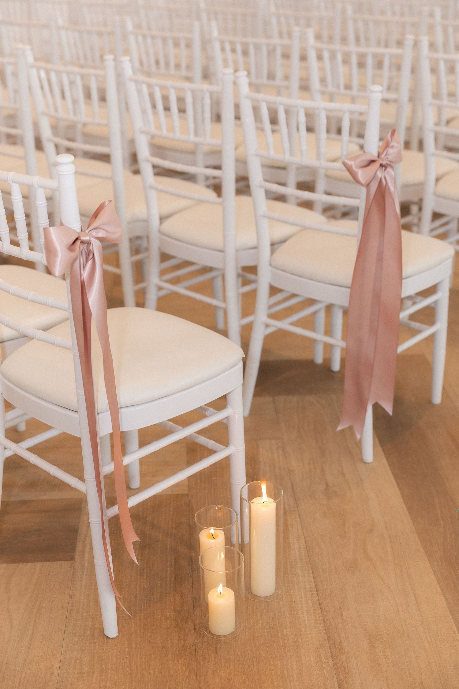 White Chiavari chairs with blush pink ribbon bows and pillar candles lining the aisle at an elegant wedding ceremony.