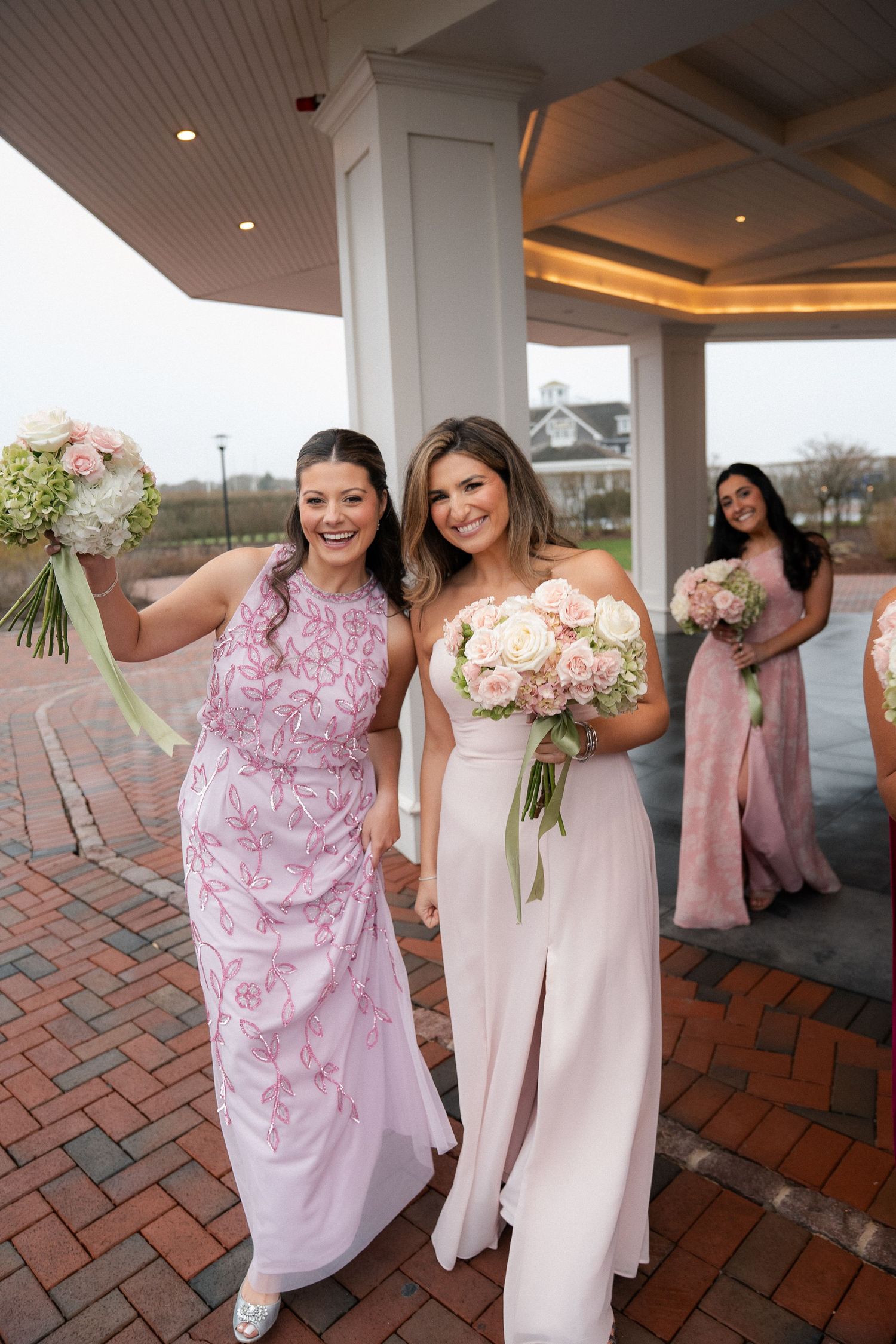 Bridesmaids in pink gowns holding floral bouquets smiling outside wedding venue pavilion.