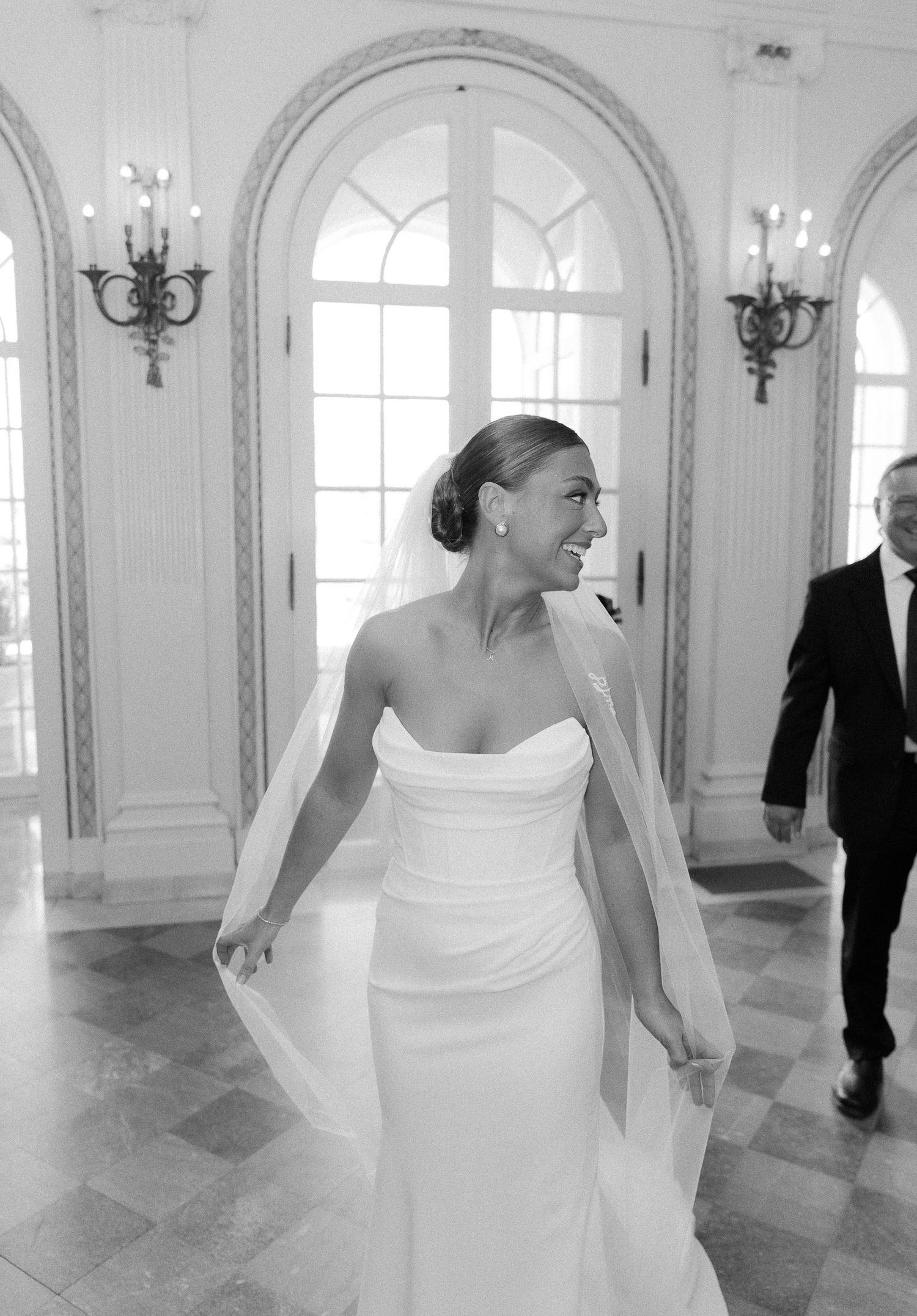 Bride in strapless white gown and veil smiles in elegant ballroom, black and white wedding photo.