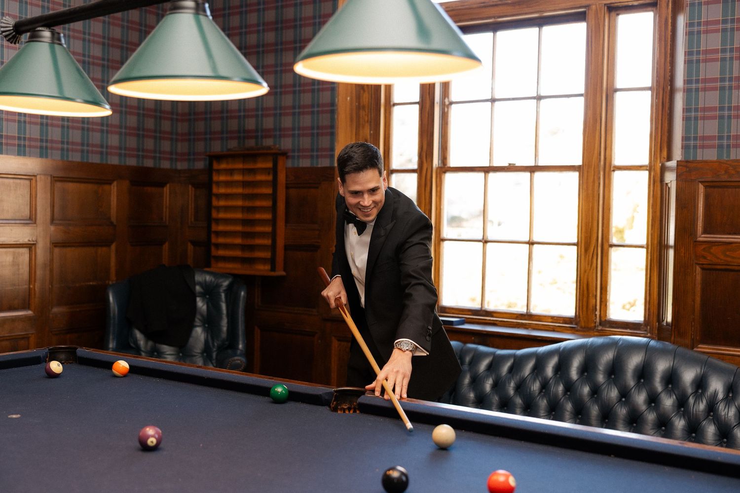 Man in suit playing pool on a billiard table in an elegant wood-paneled room with hanging lamps.
