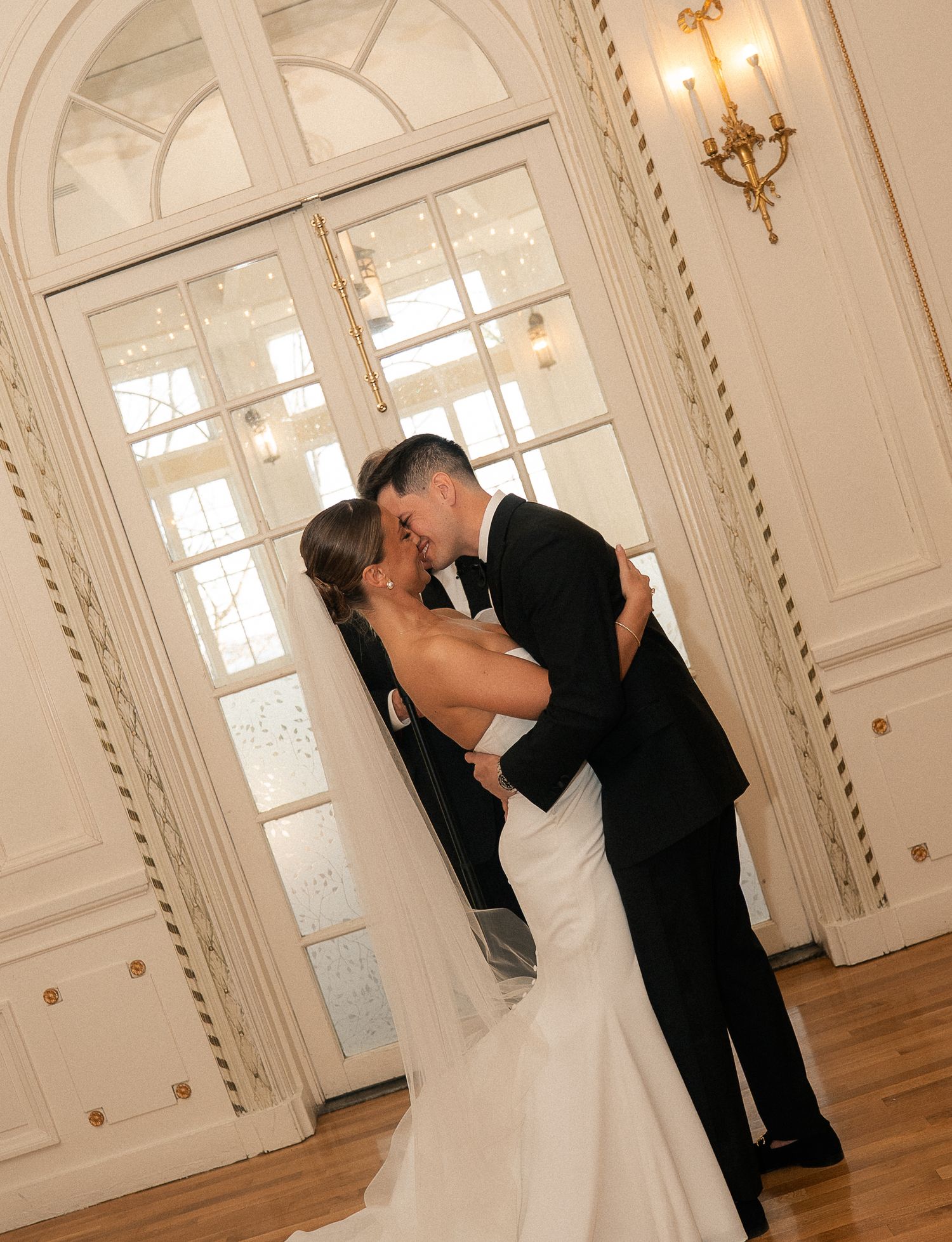 A bride and groom share a romantic kiss in an elegant ballroom, with the bride in a white gown and groom in a black tuxedo.