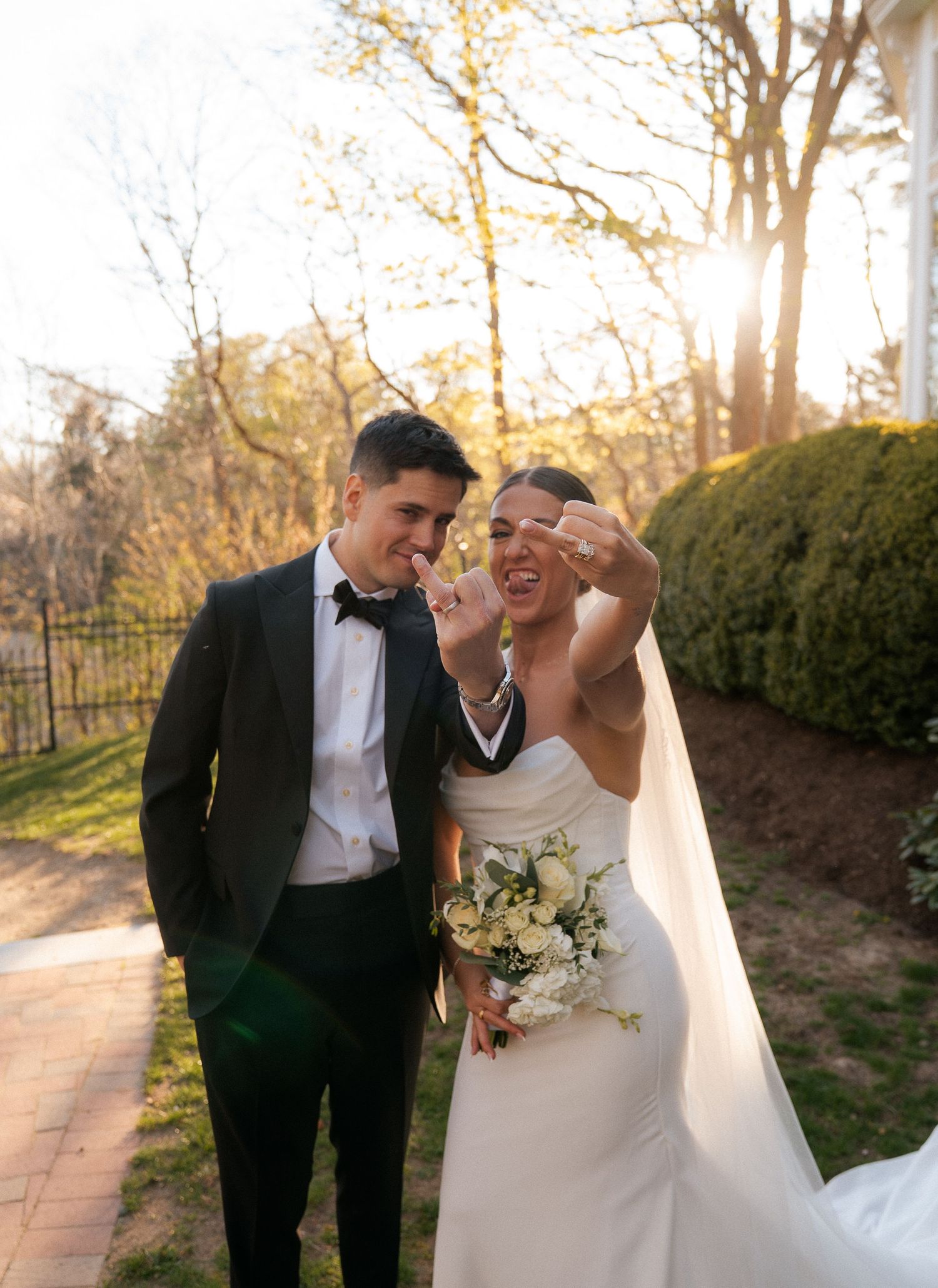 Happy newlyweds laughing outdoors at golden hour, bride in white gown showing ring, groom in black tuxedo.
