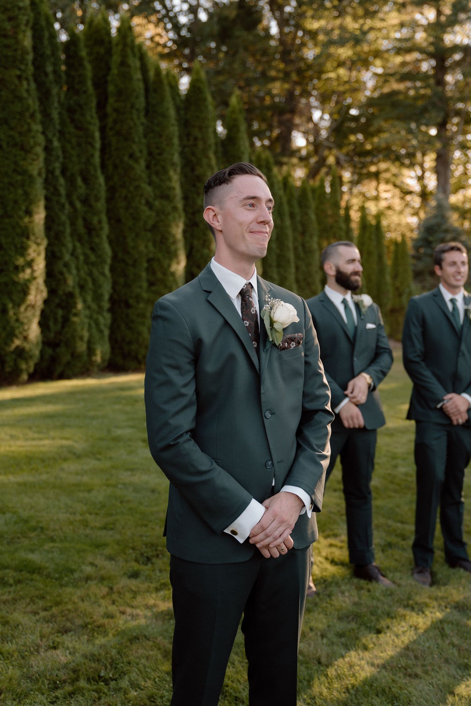Groomsmen in formal attire stand in a row against evergreen trees during sunset.