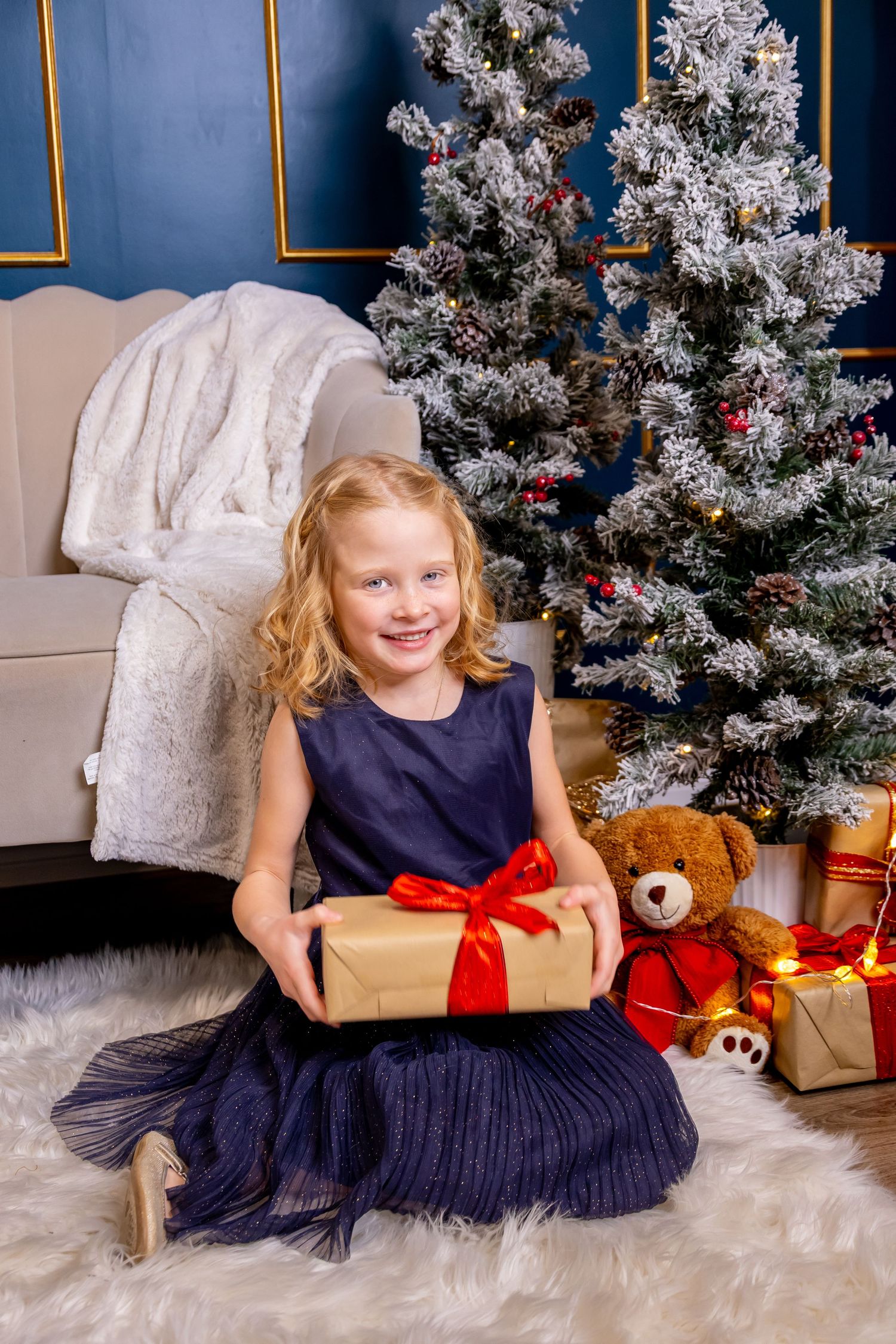 Smiling blonde girl in blue dress holding gift box with red bow sitting by decorated Christmas tree.