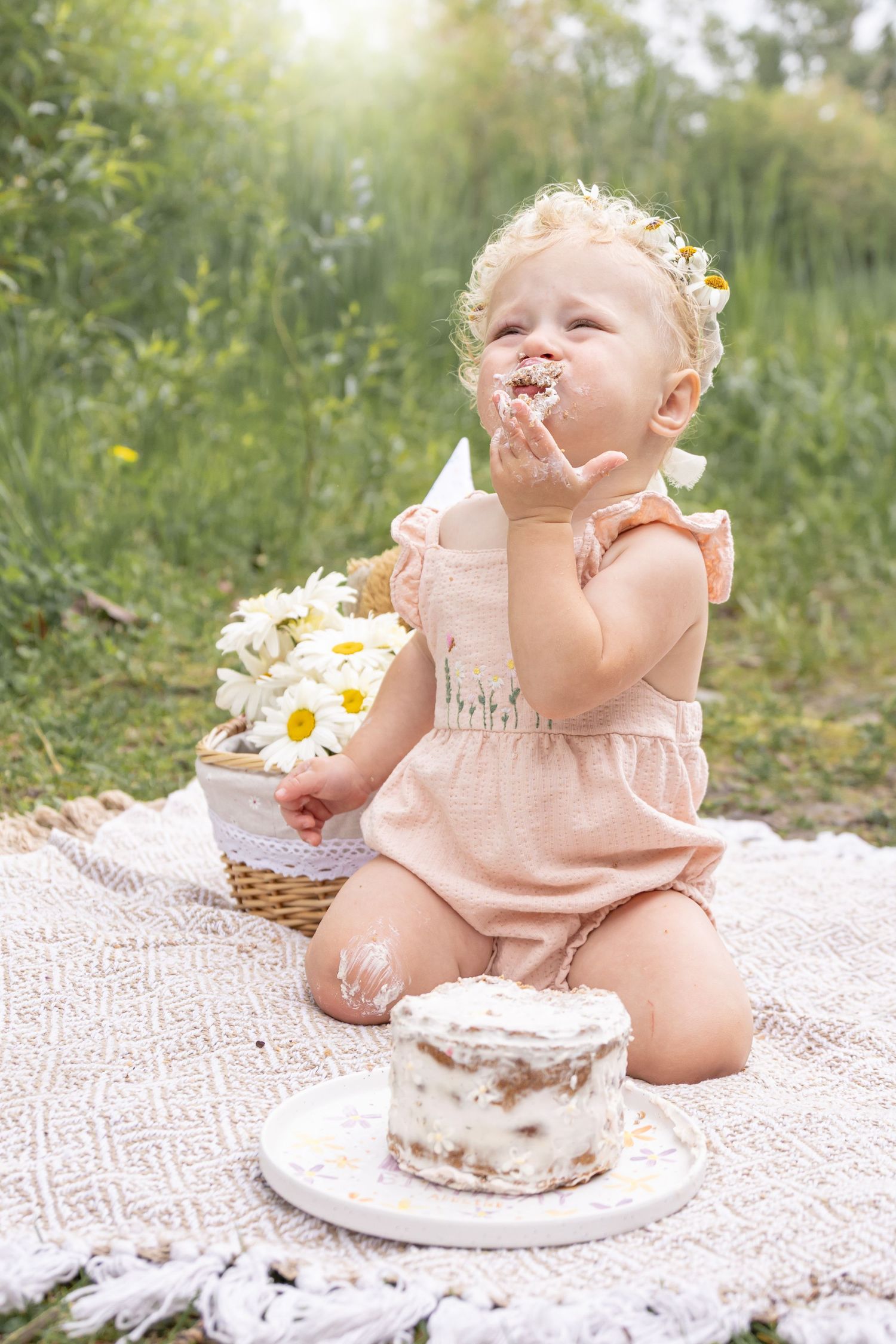 Baby in a pink romper and floral headband eats a smash cake on a woven blanket in a sunny meadow beside a basket of daisies.