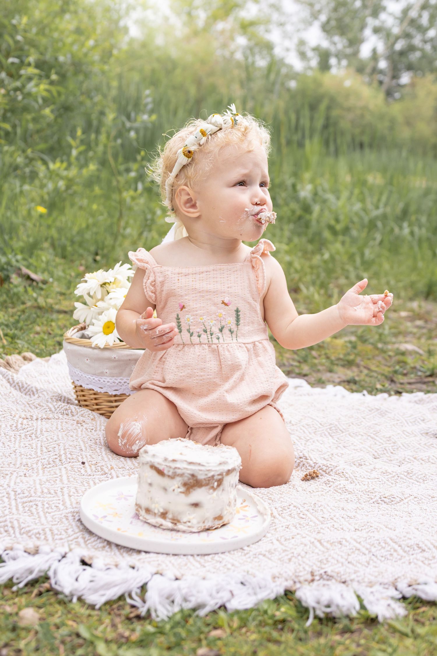 Toddler in a pink romper and flower crown enjoys a smash cake on a cream blanket in a grassy field beside a basket of daisies.