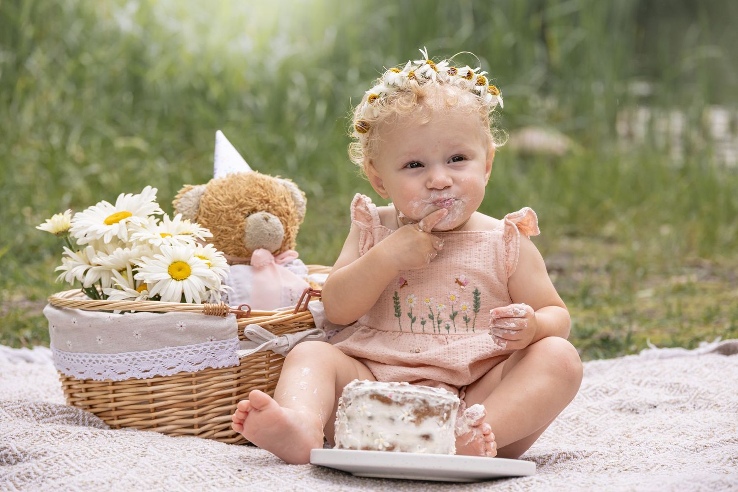 Baby girl in a floral crown and pink dress eats a small cake at a daisy picnic with a teddy bear and basket on a blanket outdoors.