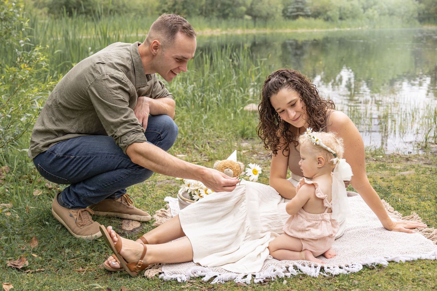 A family with a baby girl sits on a white blanket by a pond during an outdoor photo session.